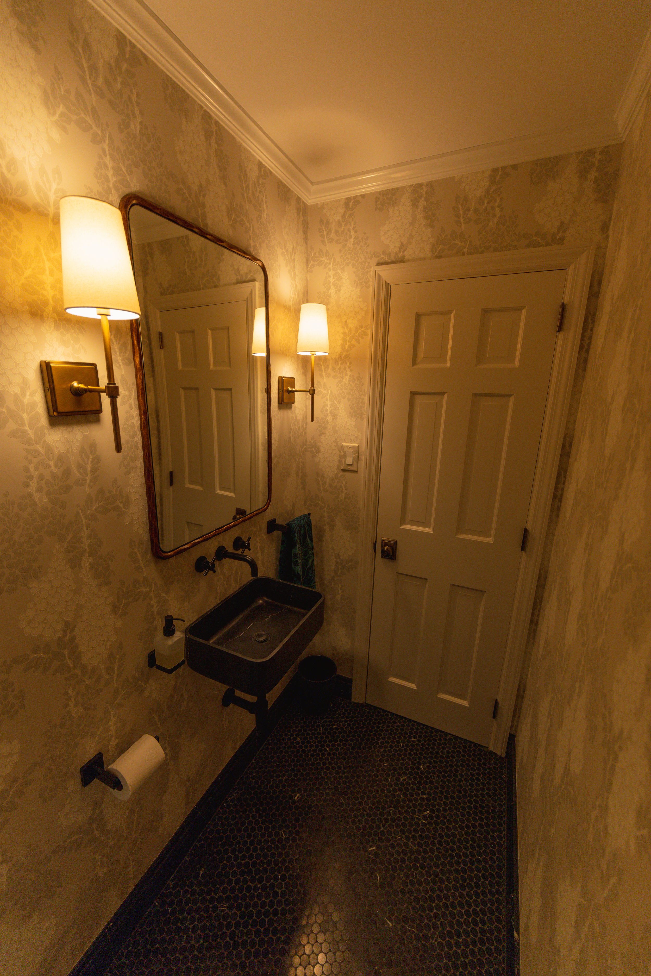 A small vintage-style bathroom with floral wallpaper, a black sink, a large mirror, two wall sconces, and a closed 6-panel door.