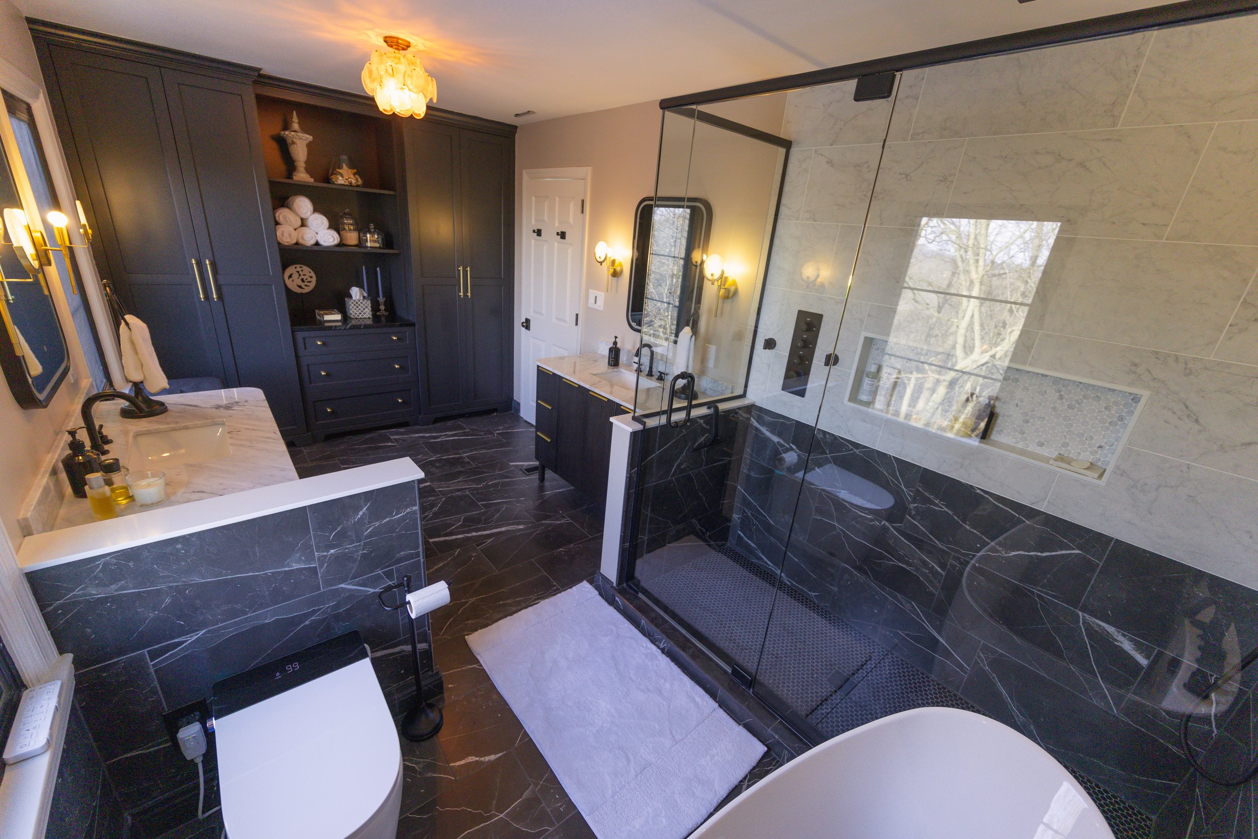Modern bathroom with black marble flooring, navy blue cabinets, a glass shower enclosure, a white freestanding bathtub, and a white marble countertop with black fixtures.