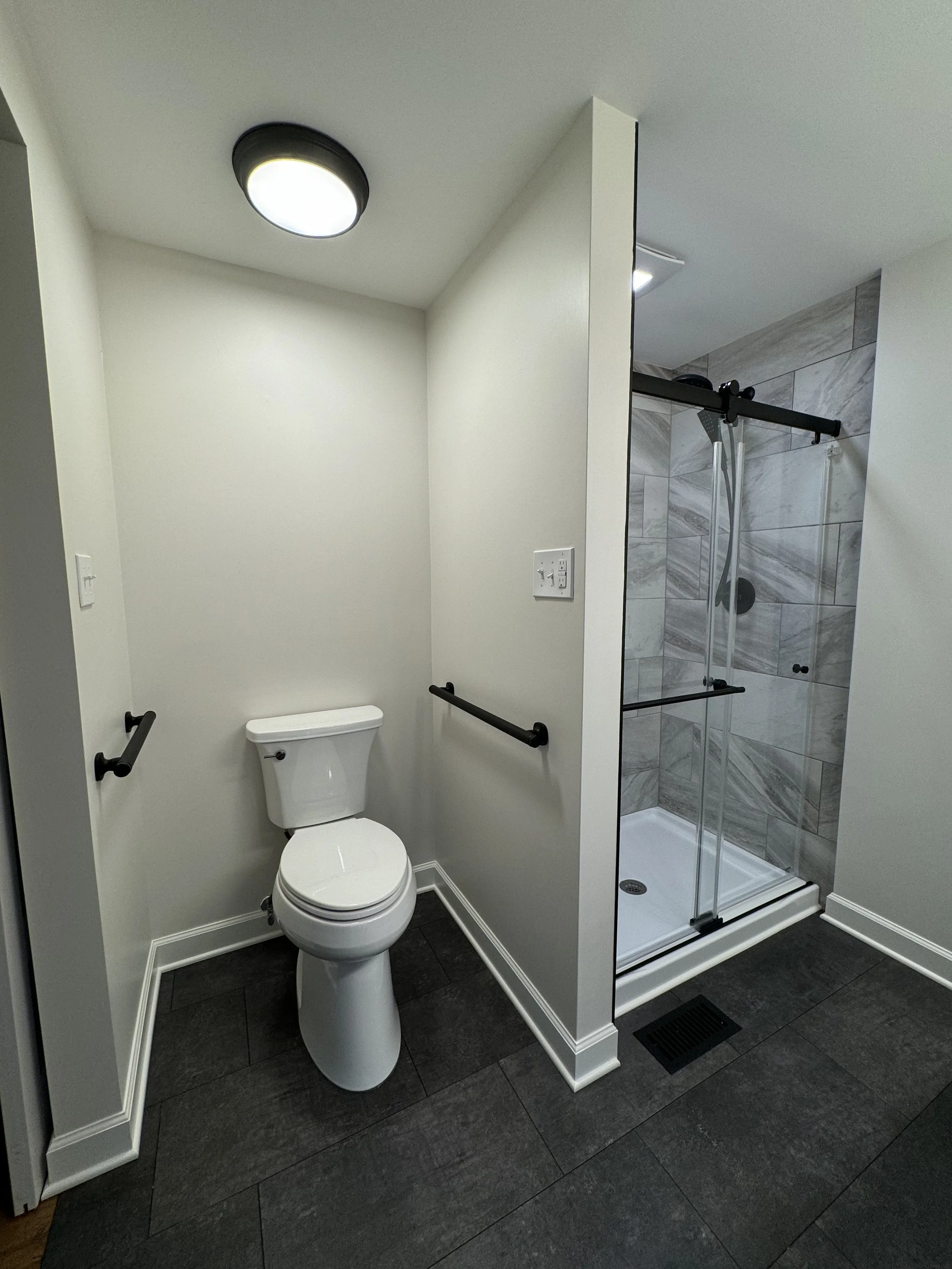 A bathroom featuring a white toilet with black grab bars on the wall, a small white shower with sliding glass doors, gray marble tiles inside the shower, black floor tiles, a ceiling light, and a wall outlet.