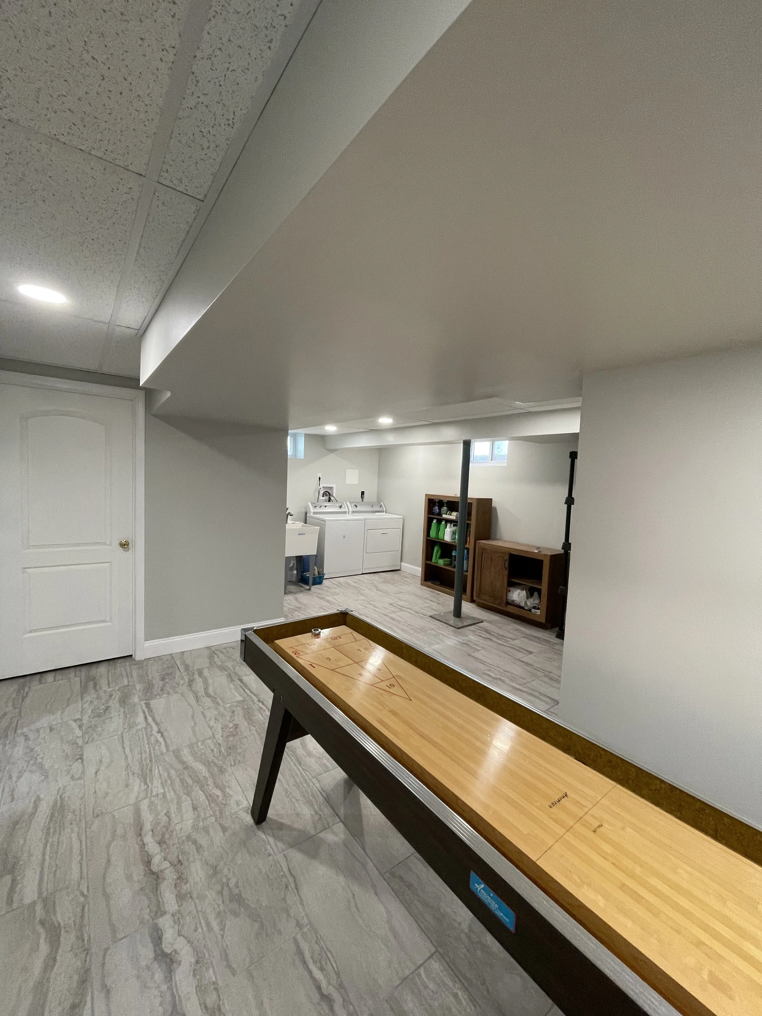 Basement recreation room with an air hockey table, washer and dryer, wood shelving, and storage cabinets, with tile flooring and recessed lighting.