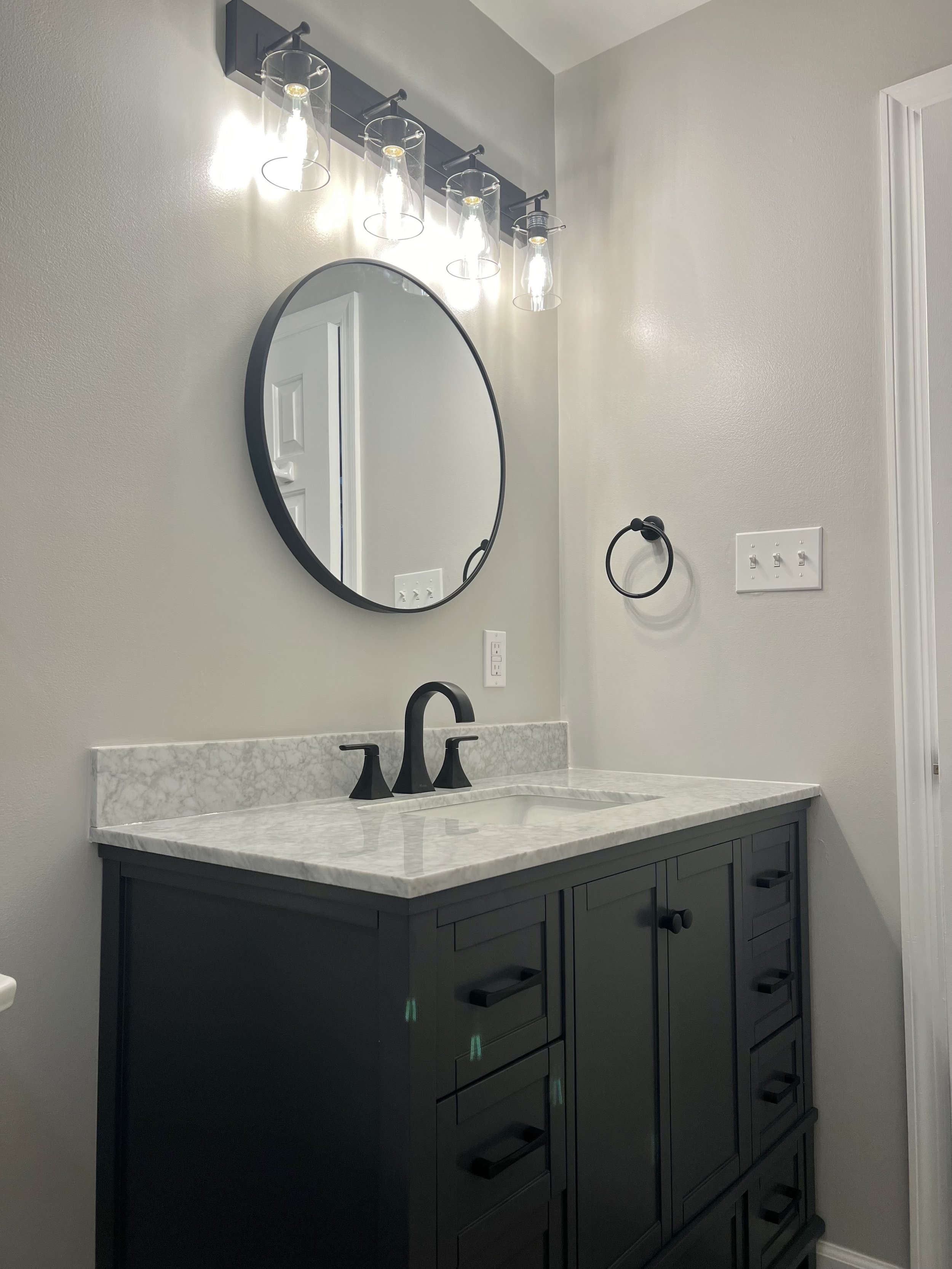 Modern bathroom vanity with a black cabinet, white marble countertop, black faucet, oval mirror, wall-mounted light fixture with multiple bulbs, and a wall-mounted towel ring.