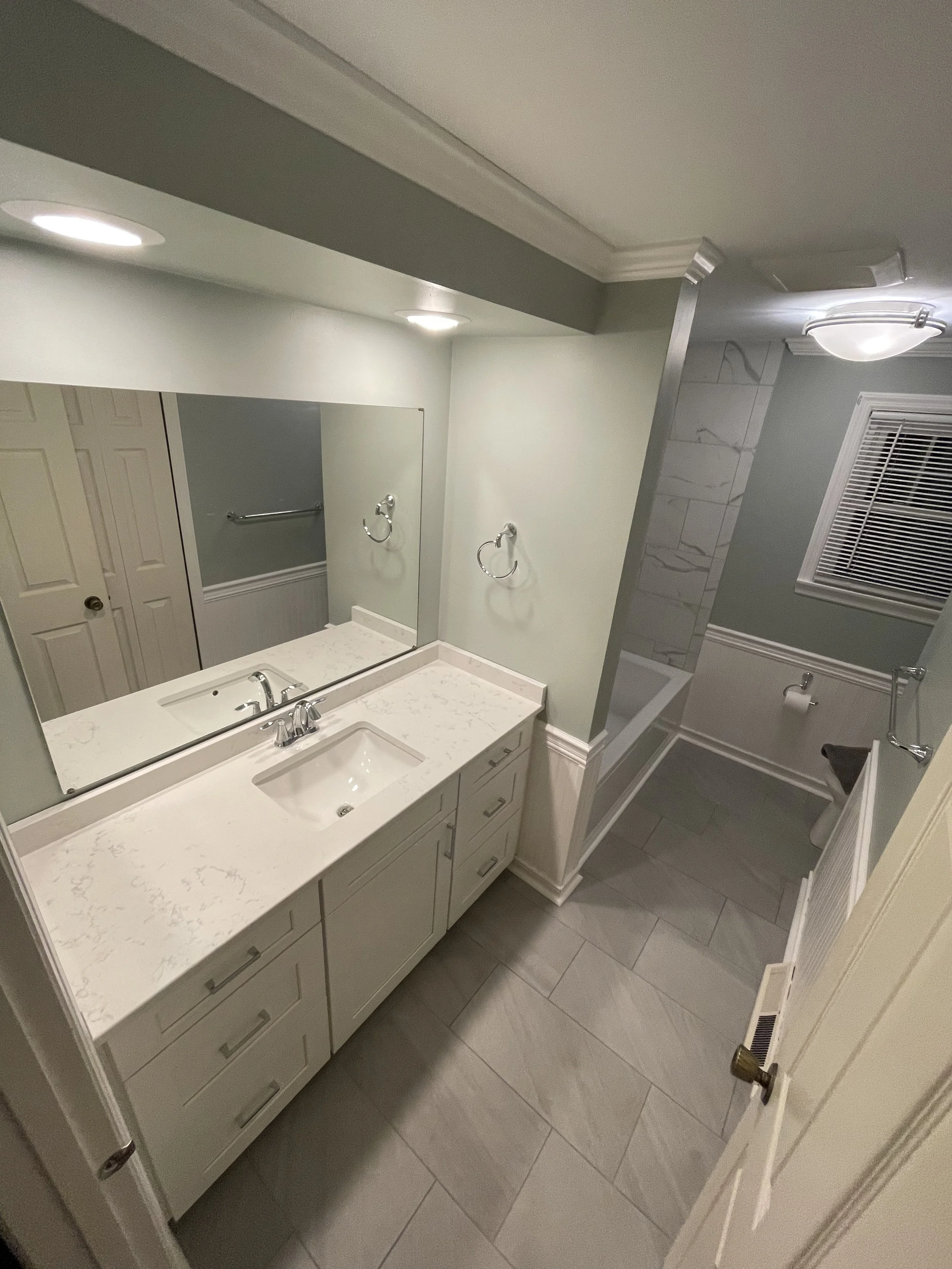 A bathroom with a white vanity, marble countertop, and an under-mount sink. A large mirror is above the vanity. There is a separate area with a bathtub, tile walls, and a window with blinds. The bathroom has overhead lighting, light green walls, and 