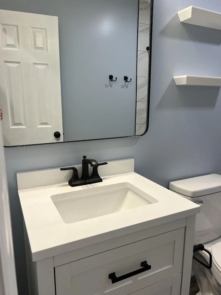 Bathroom vanity with white sink, black faucet, large mirror, light blue walls, two white shelves, and a toilet with white tank.