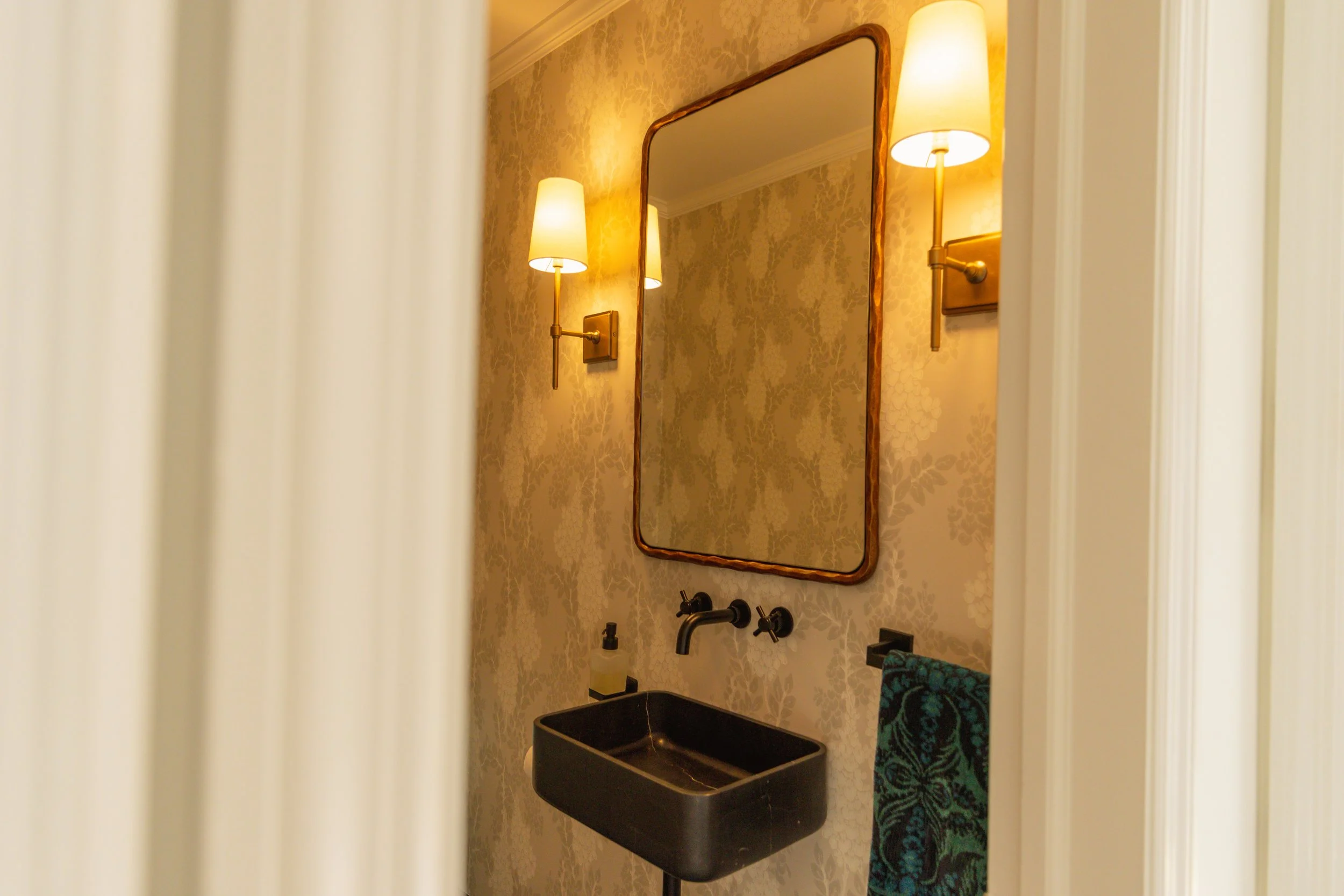 A small bathroom with beige floral wallpaper, a black sink, a mirror with a wooden frame, and two wall-mounted lamps with cream shades.