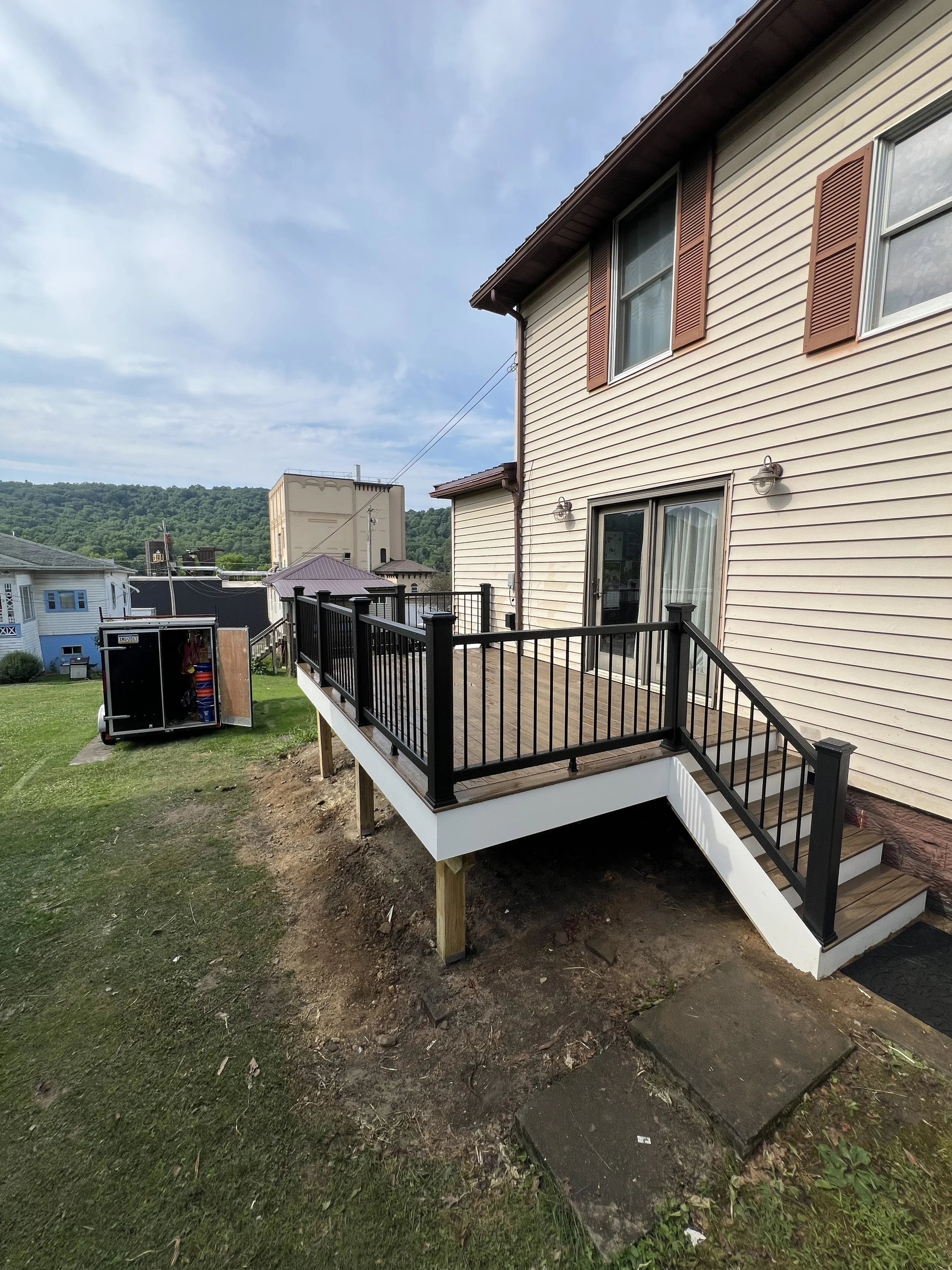 Backyard view of a house with a newly built wooden deck featuring black railing, sliding glass door, and exterior bathroom with stairs leading down.