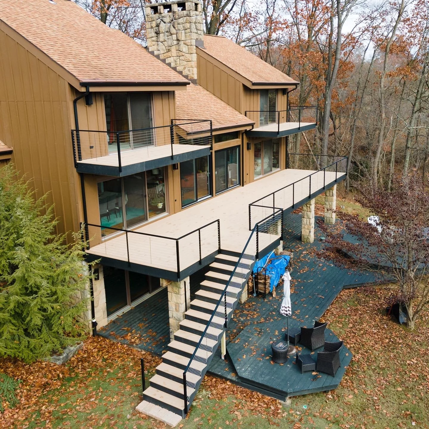 A multi-story house with a tan exterior, stone chimney, and outdoor balconies with black railings, surrounded by trees with fall foliage and fallen leaves on the ground.