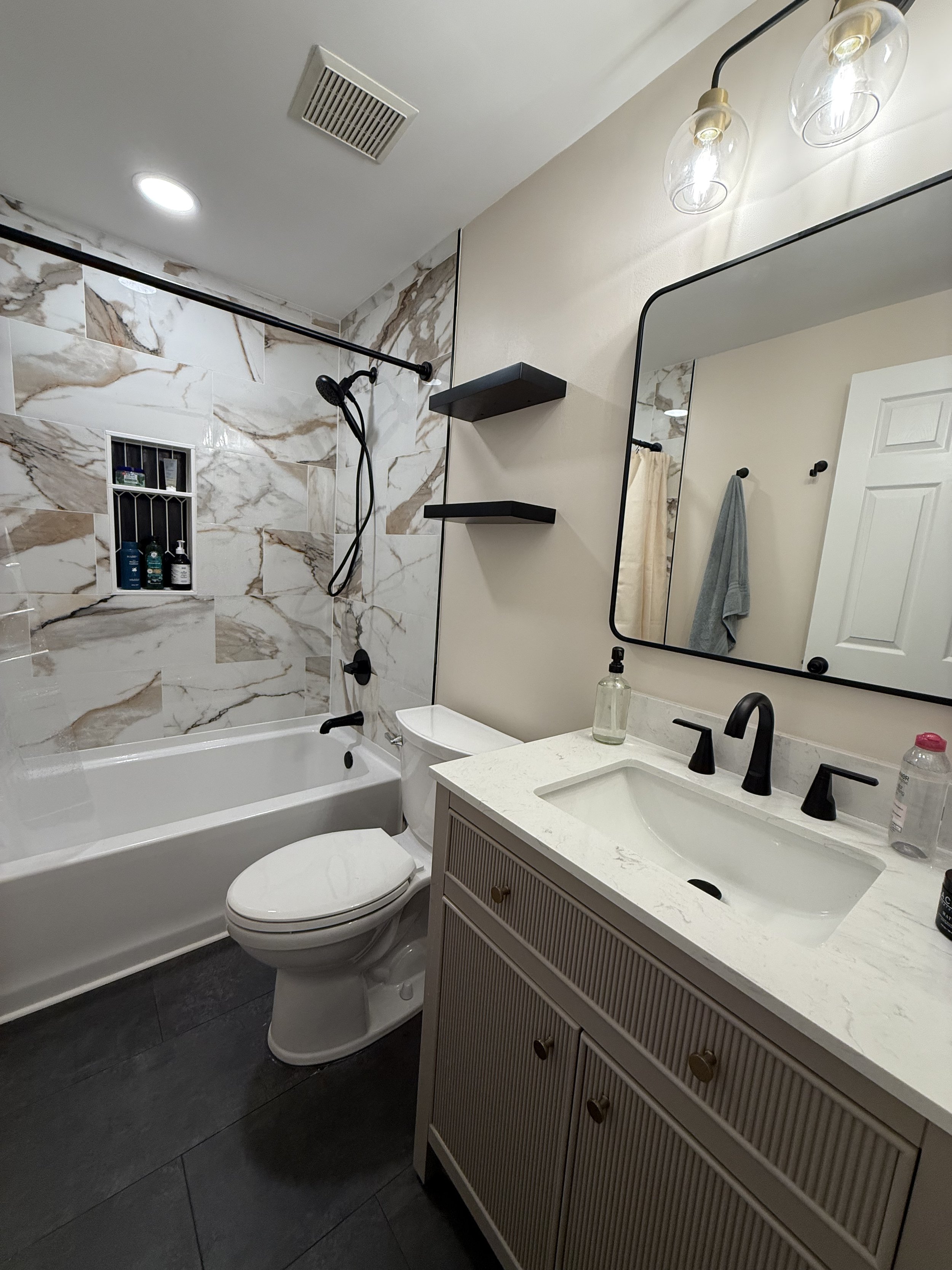 Modern bathroom with a white bathtub, marble wall tiles, a toilet, a beige vanity with a white countertop, a black faucet, and a mirror. There are black floating shelves, a beige towel, and two black light fixtures above the mirror.
