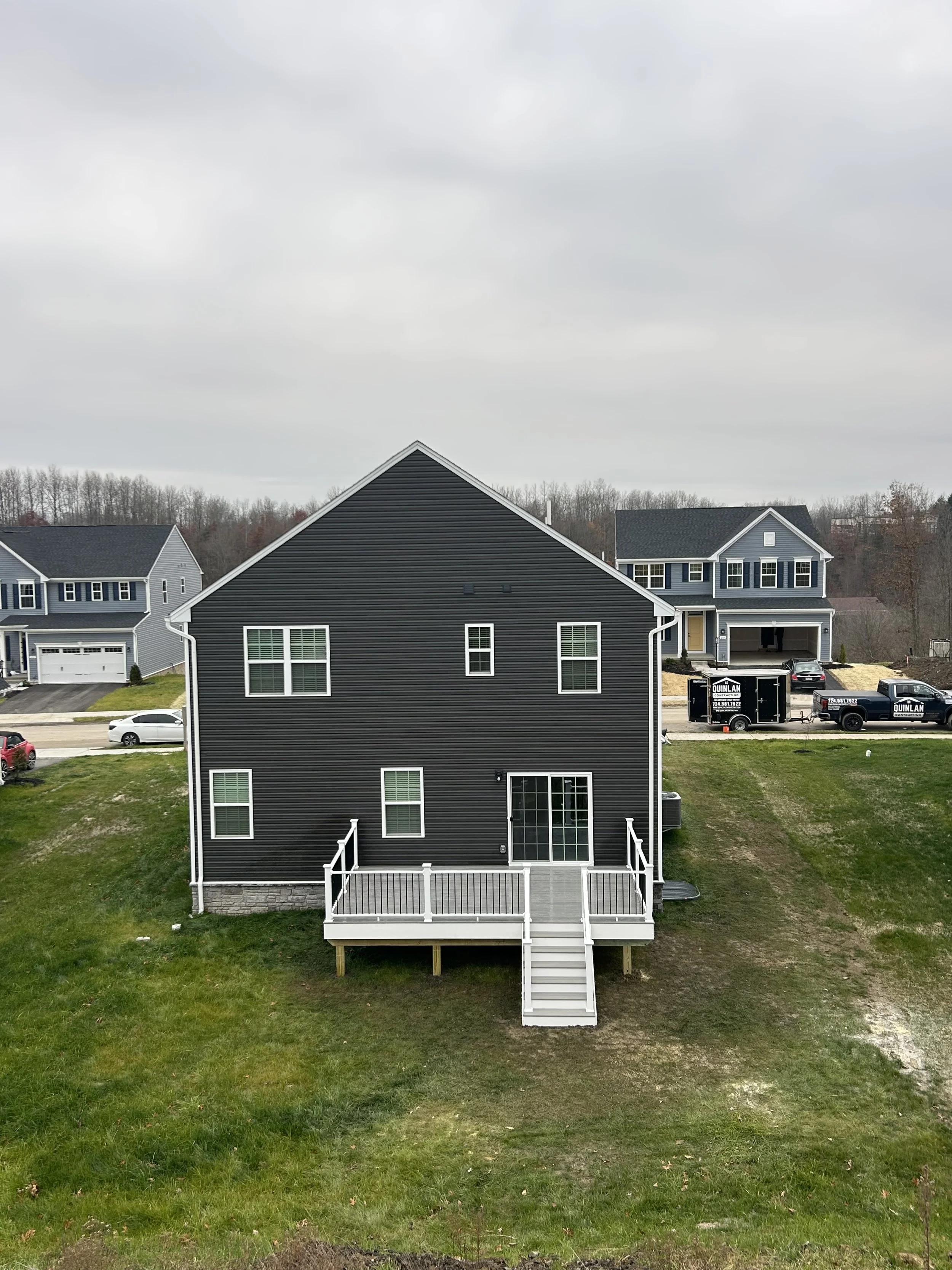 Rear view of a two-story house with dark siding, white trim, and a small deck with stairs, set on a grassy hill, with neighboring houses and trucks in the background under cloudy skies.