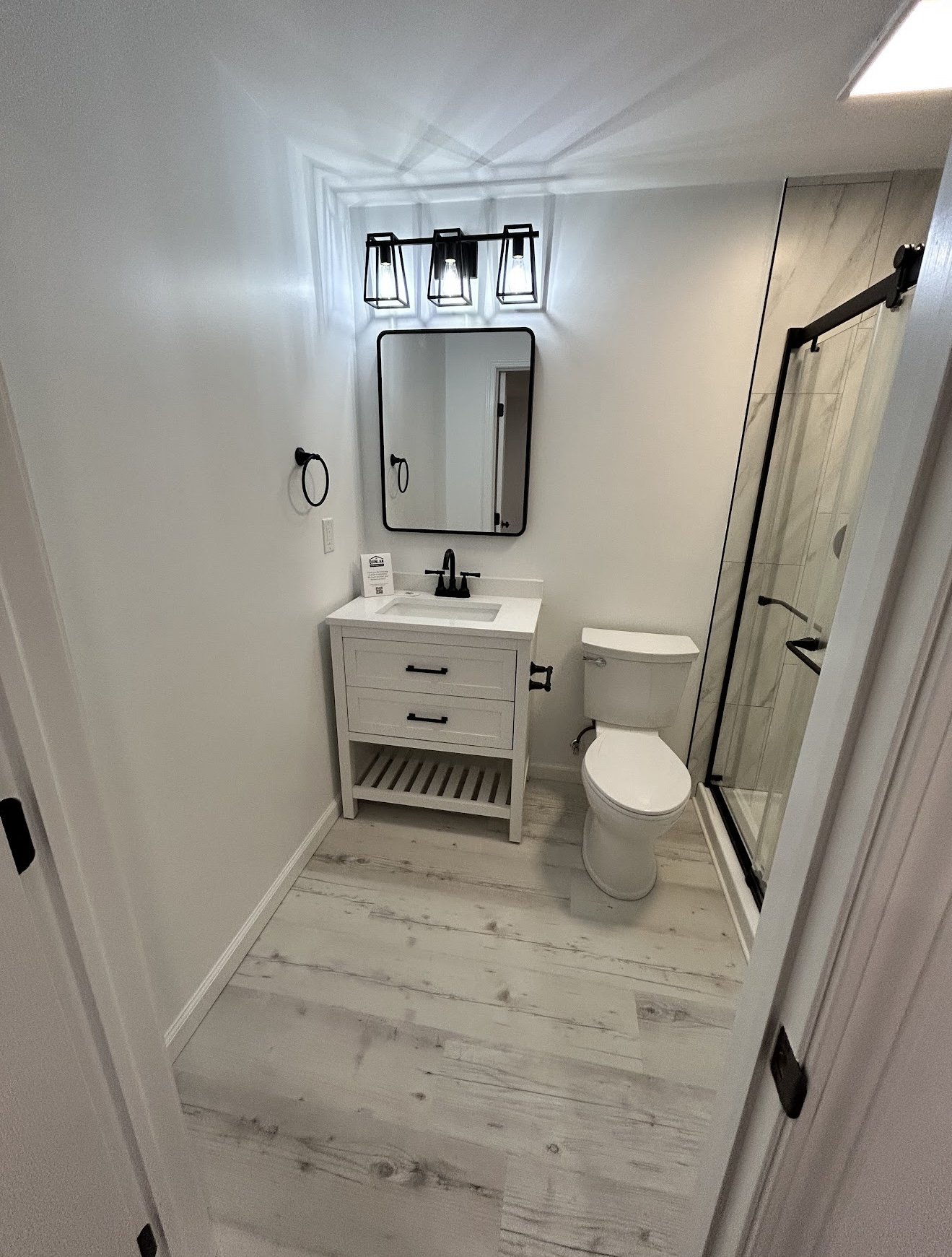 A small modern bathroom with white walls, a white vanity with drawers and open shelf, a black faucet, a rectangular mirror, black towel rings, a toilet, a glass shower door with marble-like tiles, and a black light fixture with multiple bulbs.