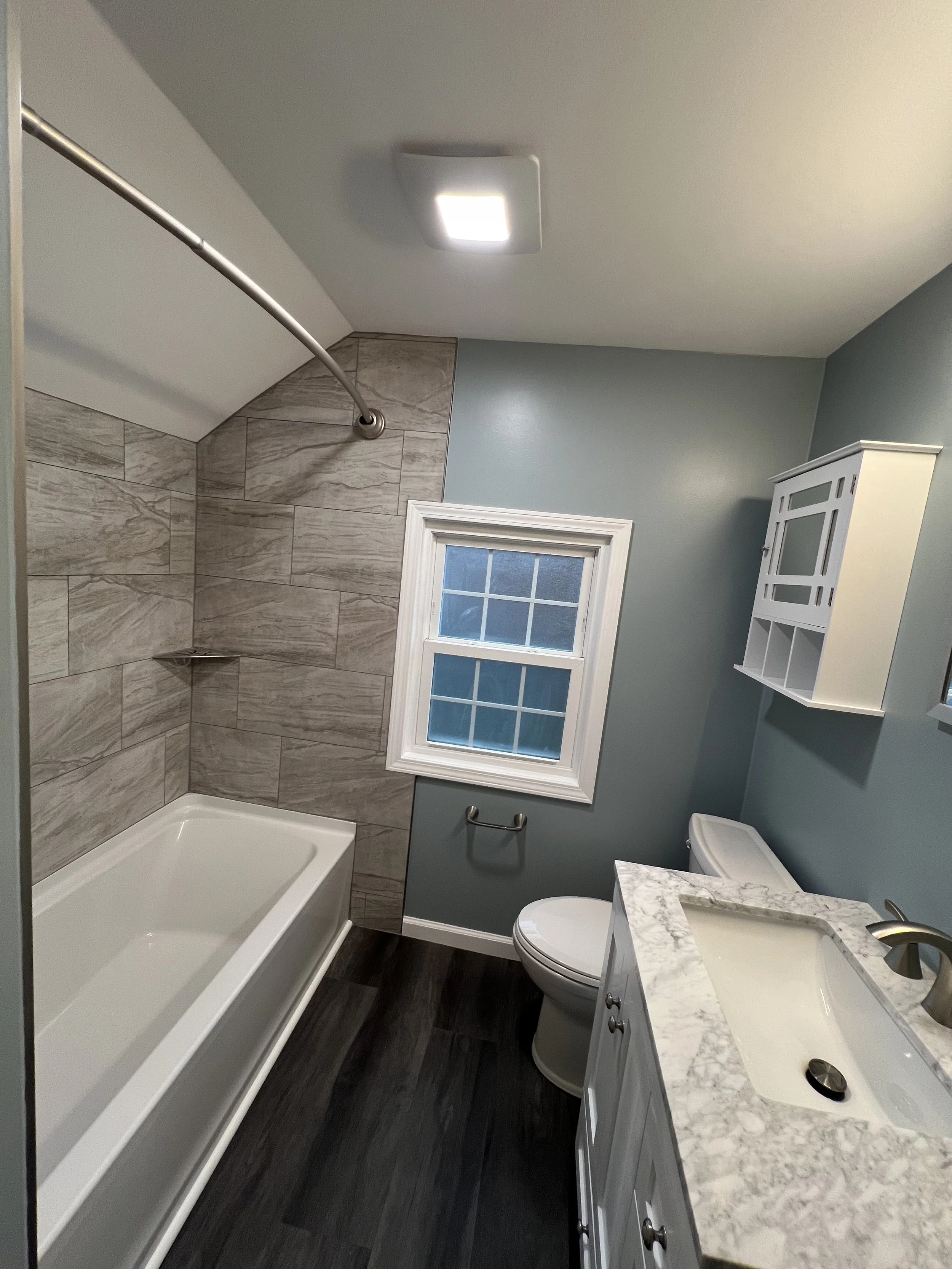 Bathroom with a bathtub, wood-look tile shower wall, frosted window, white vanity with marble countertop, undermount sink, wall-mounted cabinet, toilet, dark wood flooring, ceiling light