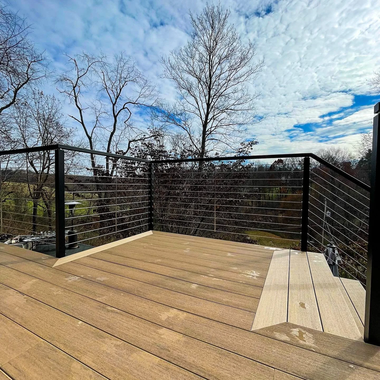 A wooden deck with metal railing, overlooking trees and a partly cloudy sky.