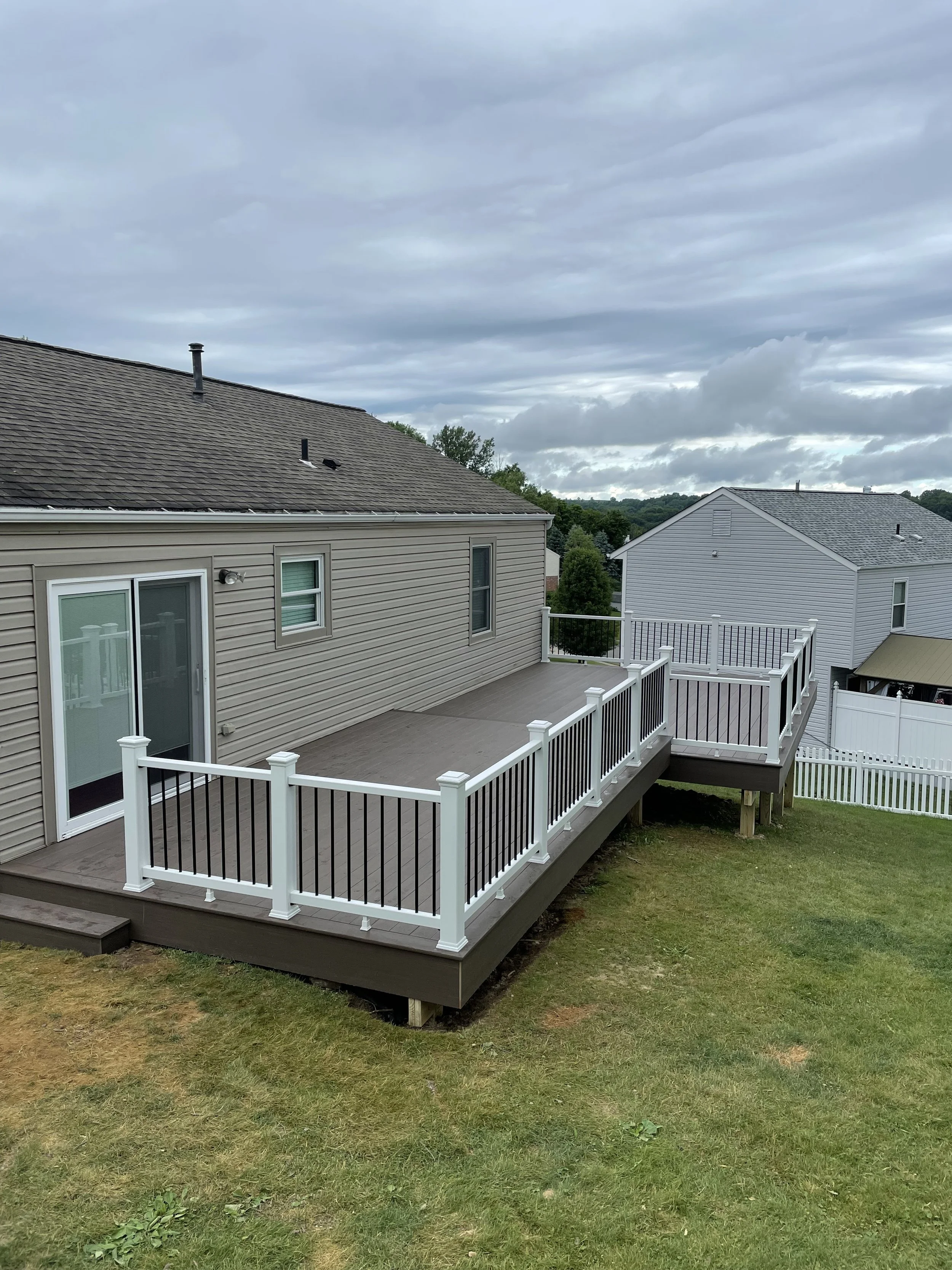 A newly built wooden deck with white and black railing around a beige house, overlooking a grassy backyard with neighboring houses and a cloudy sky.