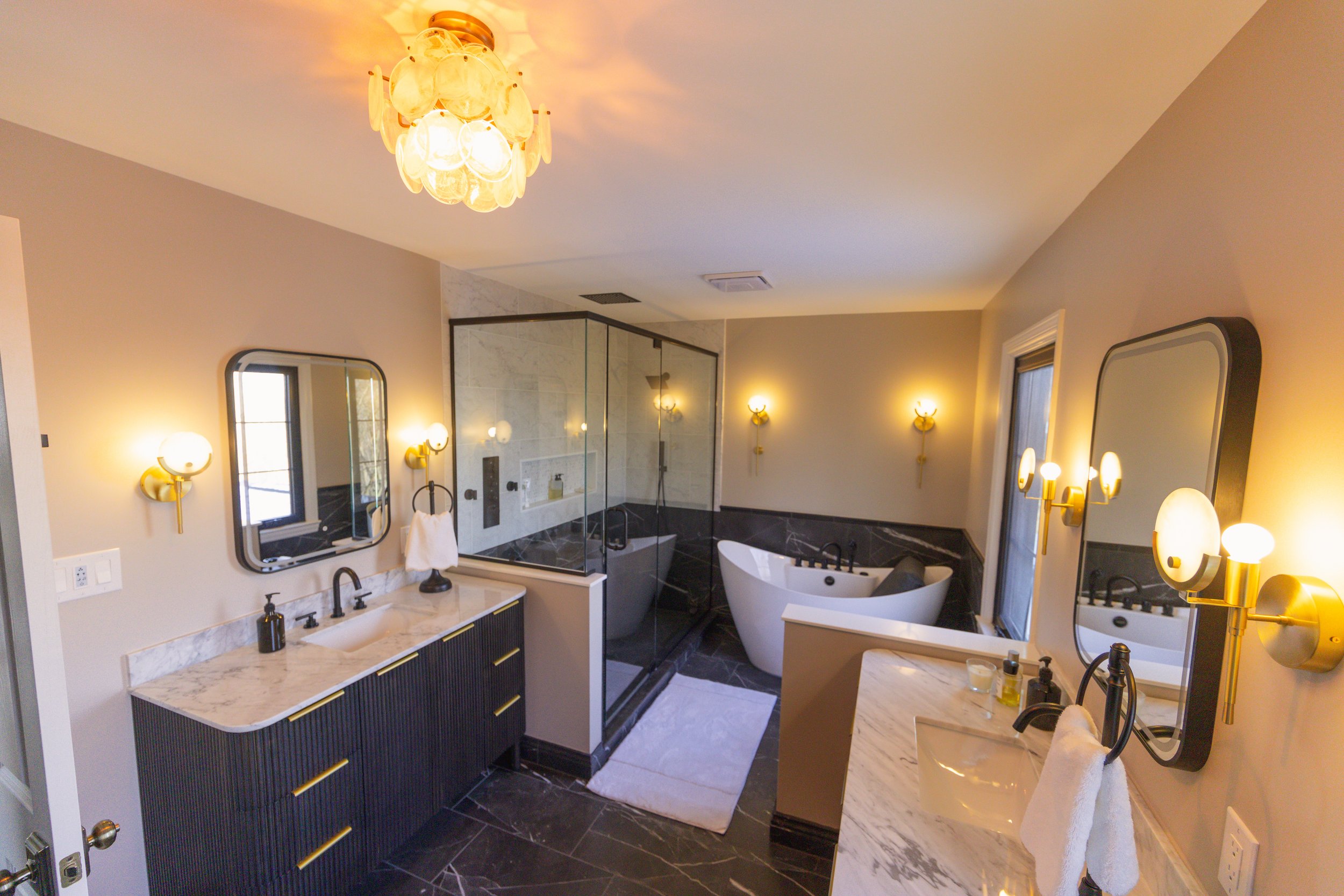 Modern bathroom with two vanities, a walk-in shower with glass doors, and a freestanding bathtub, all illuminated by warm wall-mounted lights.