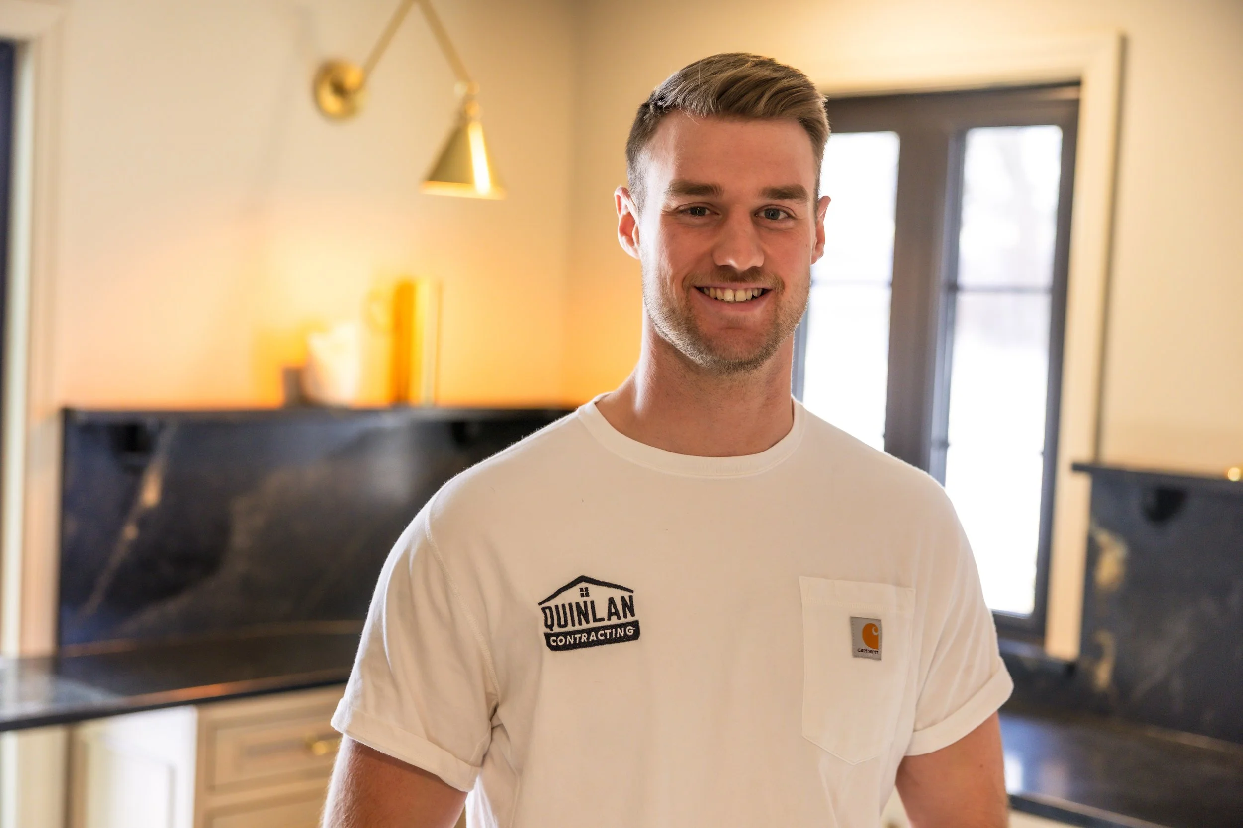 A smiling man in a white T-shirt with patched logos, standing in a kitchen with a window, black countertop, and warm lighting.