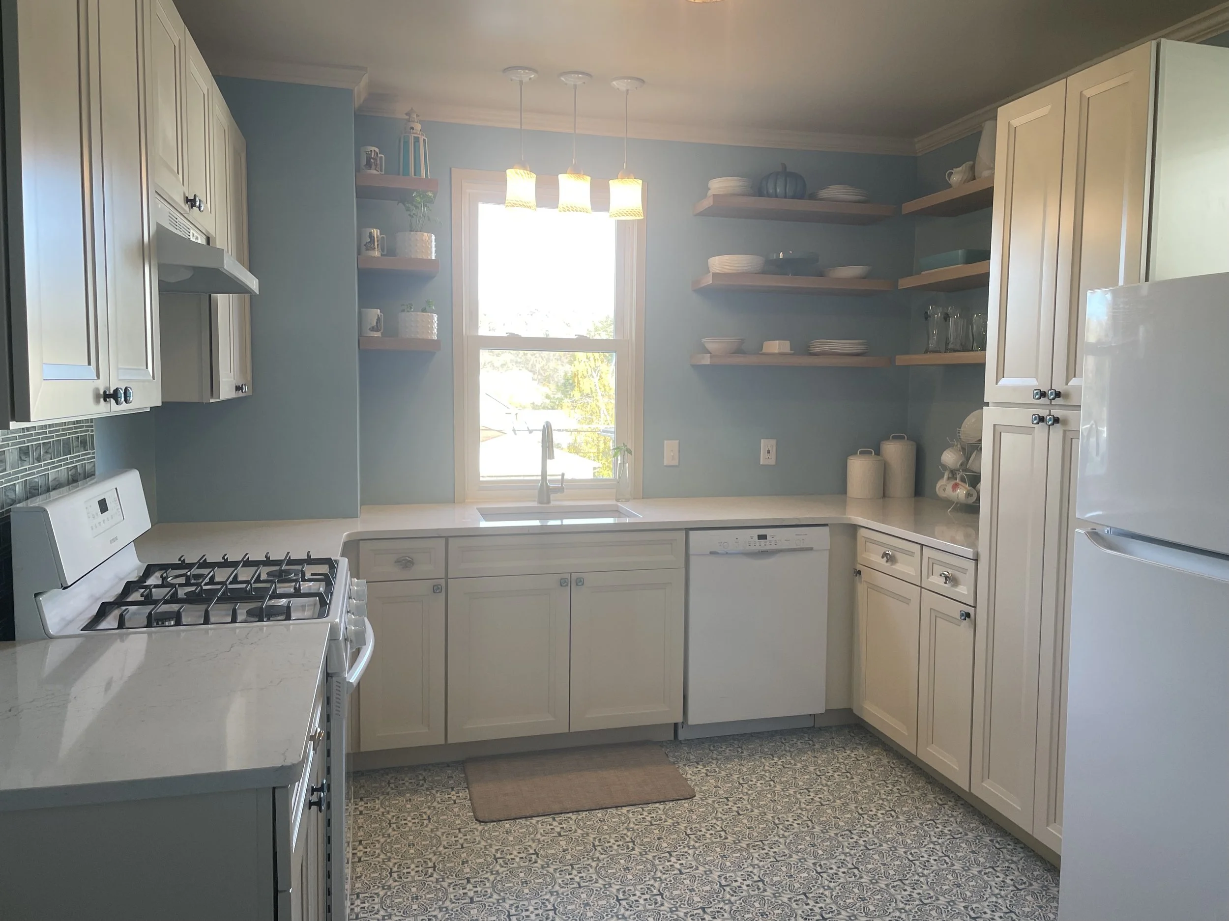 A kitchen with light-colored cabinets, open wooden shelves with dishes and glasses, a white refrigerator, dishwasher, and stove, a window above the sink, and patterned floor tiles.
