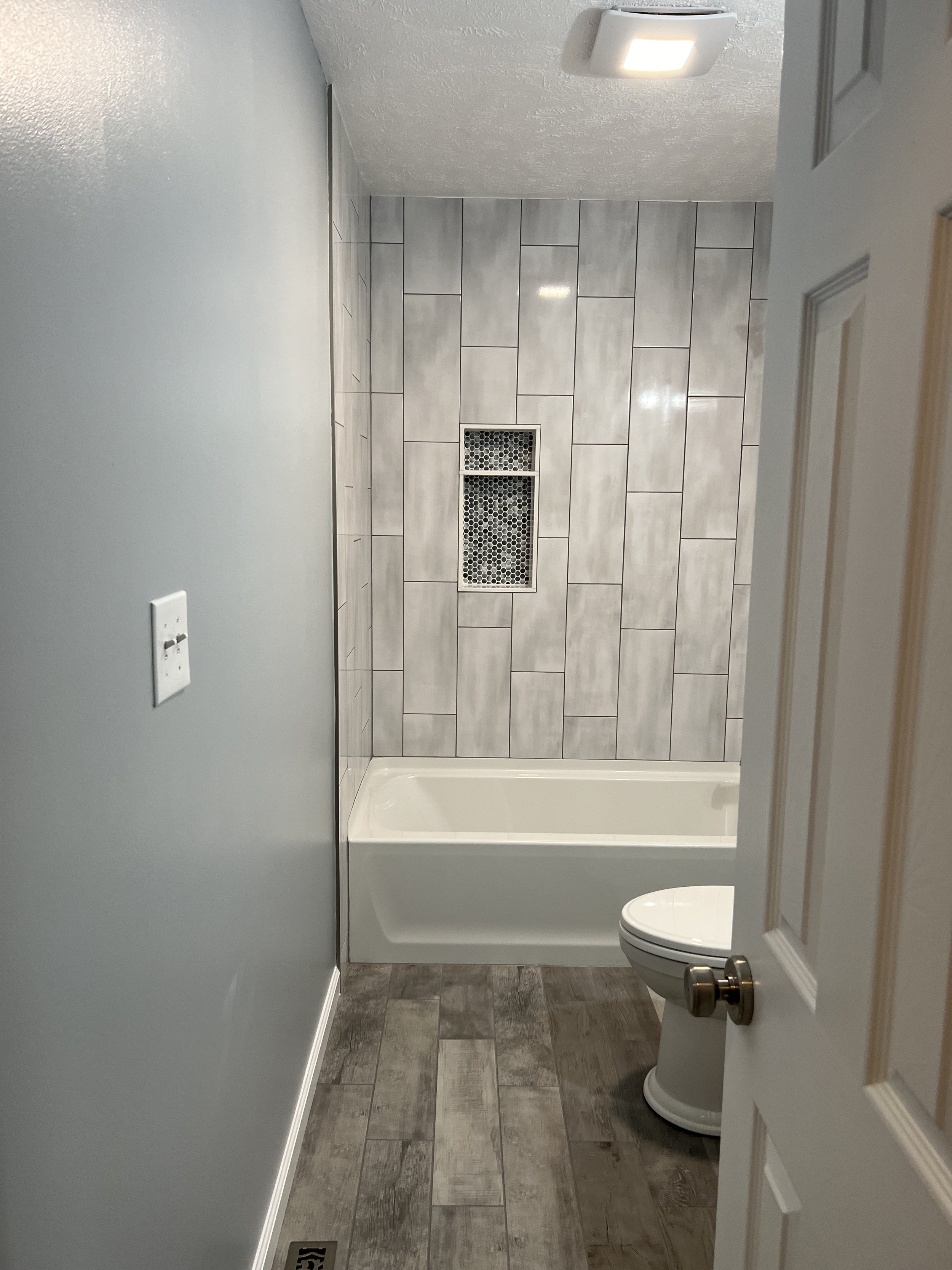 A small bathroom with a gray tiled shower wall, a white bathtub, a toilet, and gray wood-style flooring.