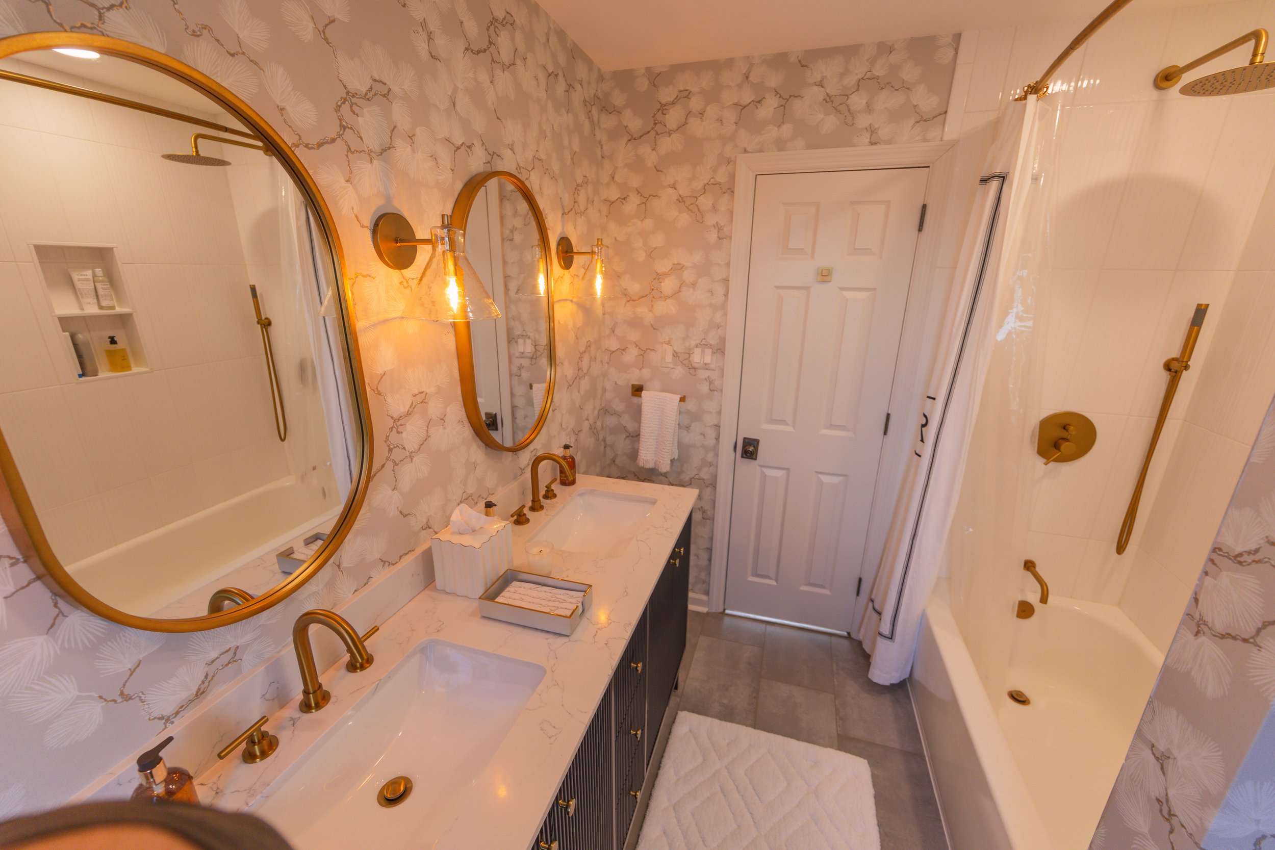 A bathroom with a double vanity sink, gold fixtures, and an oval mirror. The wall behind has a floral wallpaper, and there is a white door. To the right, there is a shower with a curtain and gold fixtures, and a white bathtub. The floor has gray tile
