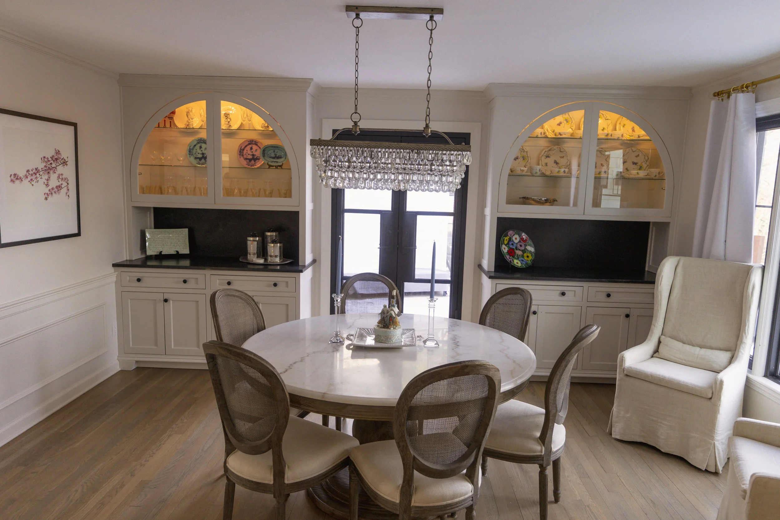 Dining room with oval marble table, six upholstered chairs, built-in china cabinets, chandelier, sliding glass doors, and white armchair.