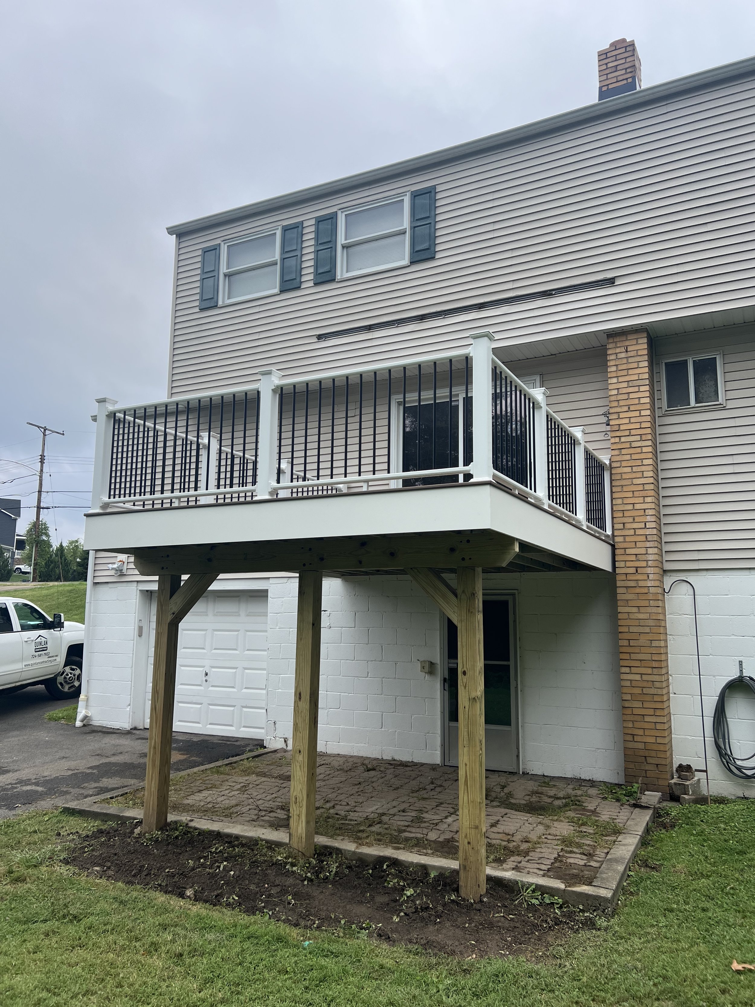 Backyard of a two-story house with a newly built wooden deck and railing. The deck has support beams and faces a patio area with bricks. The house has beige siding, a brick chimney, and windows with shutters.