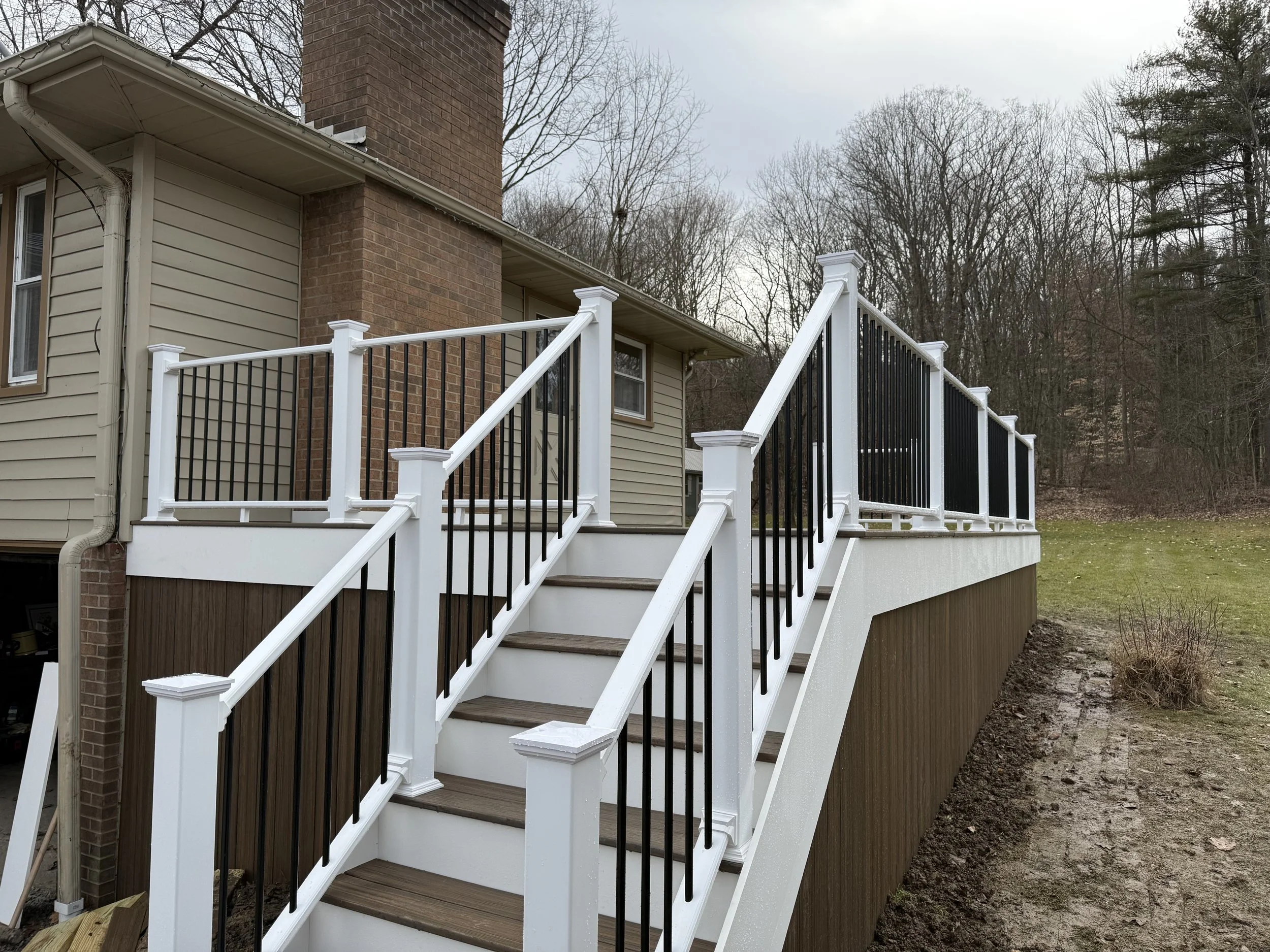 Wooden deck with white and black railing attached to a beige house with a brick chimney, with stairs leading down to grass.