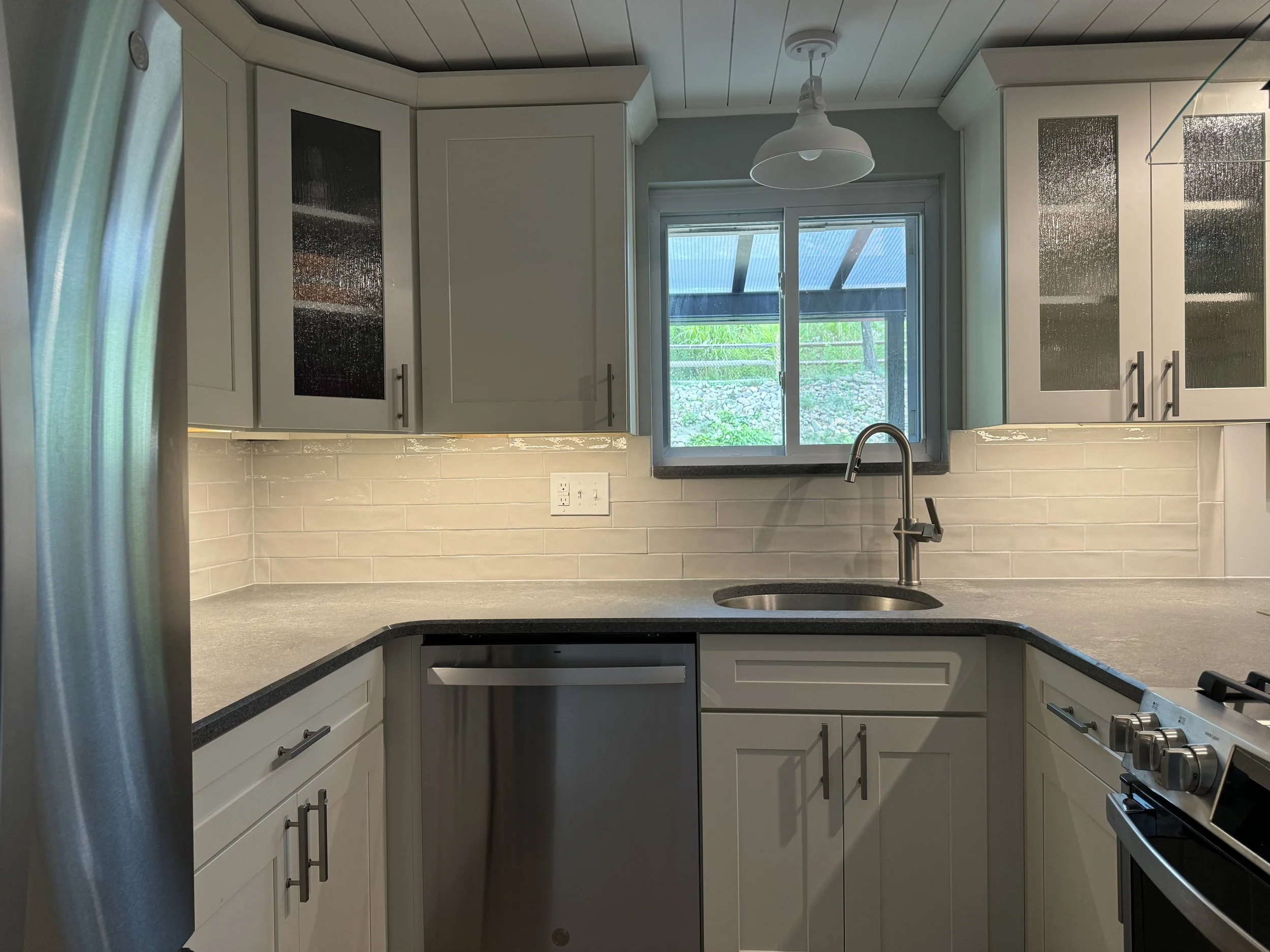 Kitchen with white cabinets, gray countertop, stainless steel appliances, beige tile backsplash, window above sink, and a white pendant light.