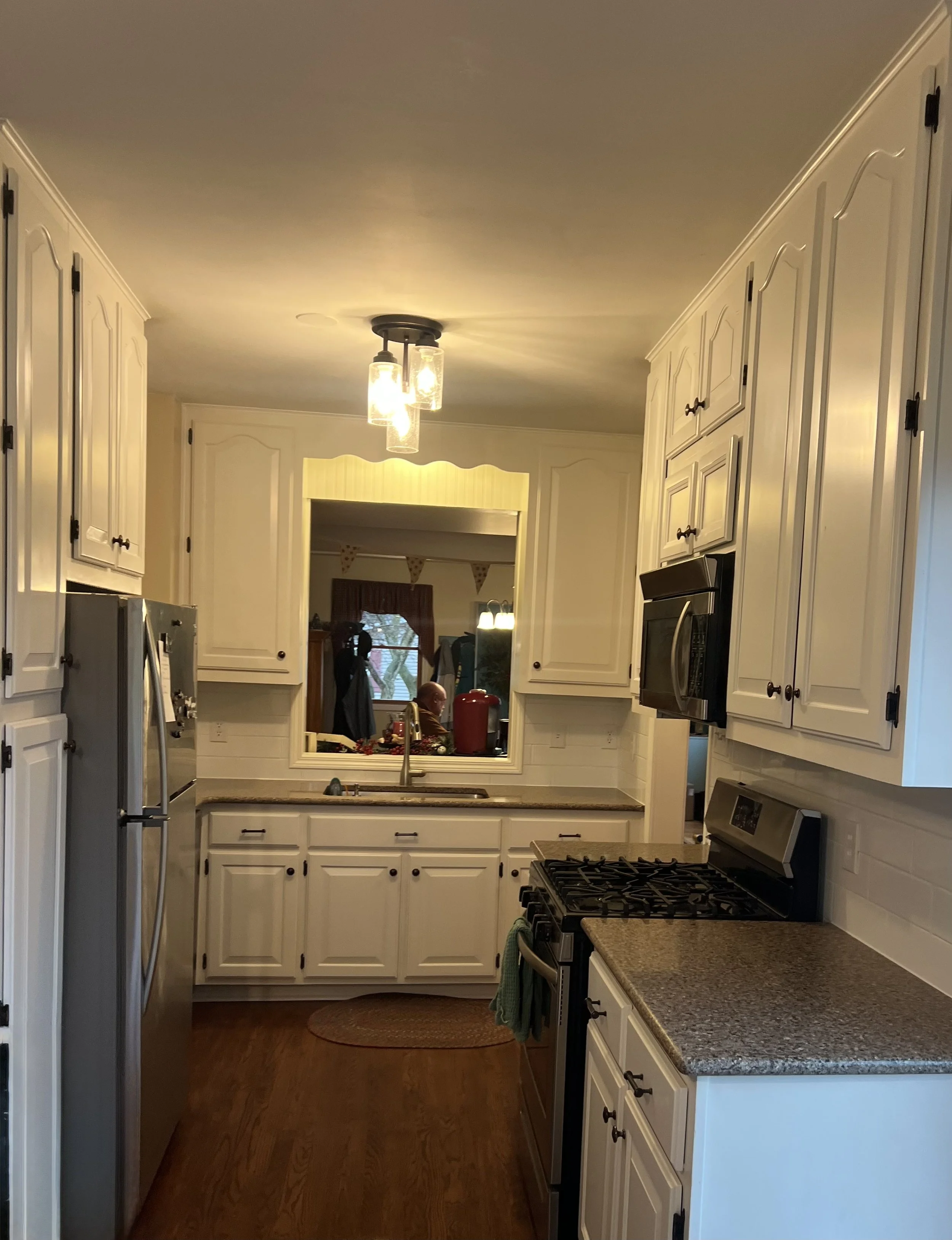 A cozy kitchen with white cabinets, a stainless steel refrigerator on the left, a black microwave above a stainless steel gas stove, beige countertops, a small window above the sink, and warm lighting, with a person visible in a reflection in the win