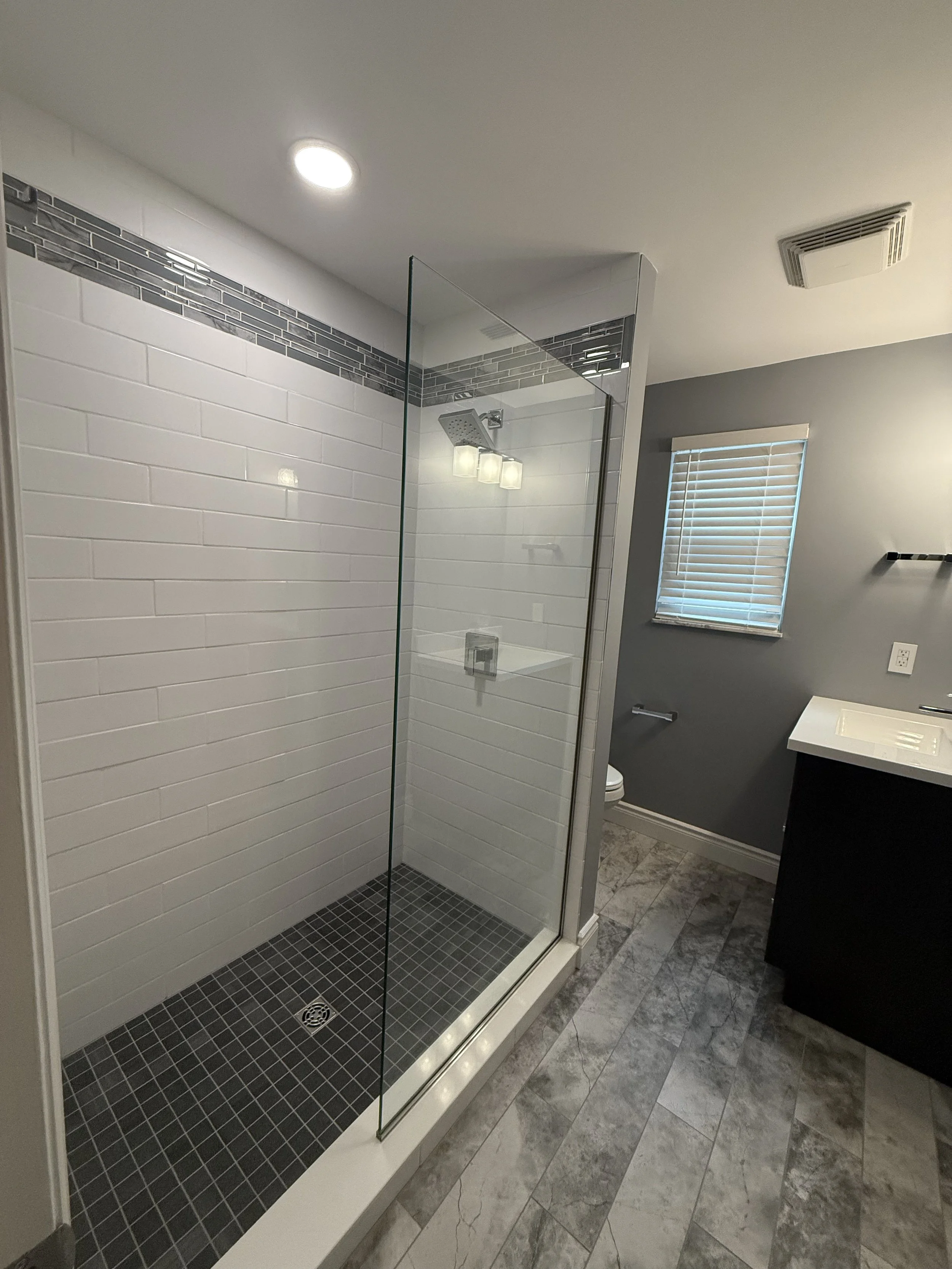 Bathroom with a glass-enclosed shower featuring white subway tiles and dark gray accent tiles, a window with blinds, a vanity with a white sink, and gray patterned flooring.