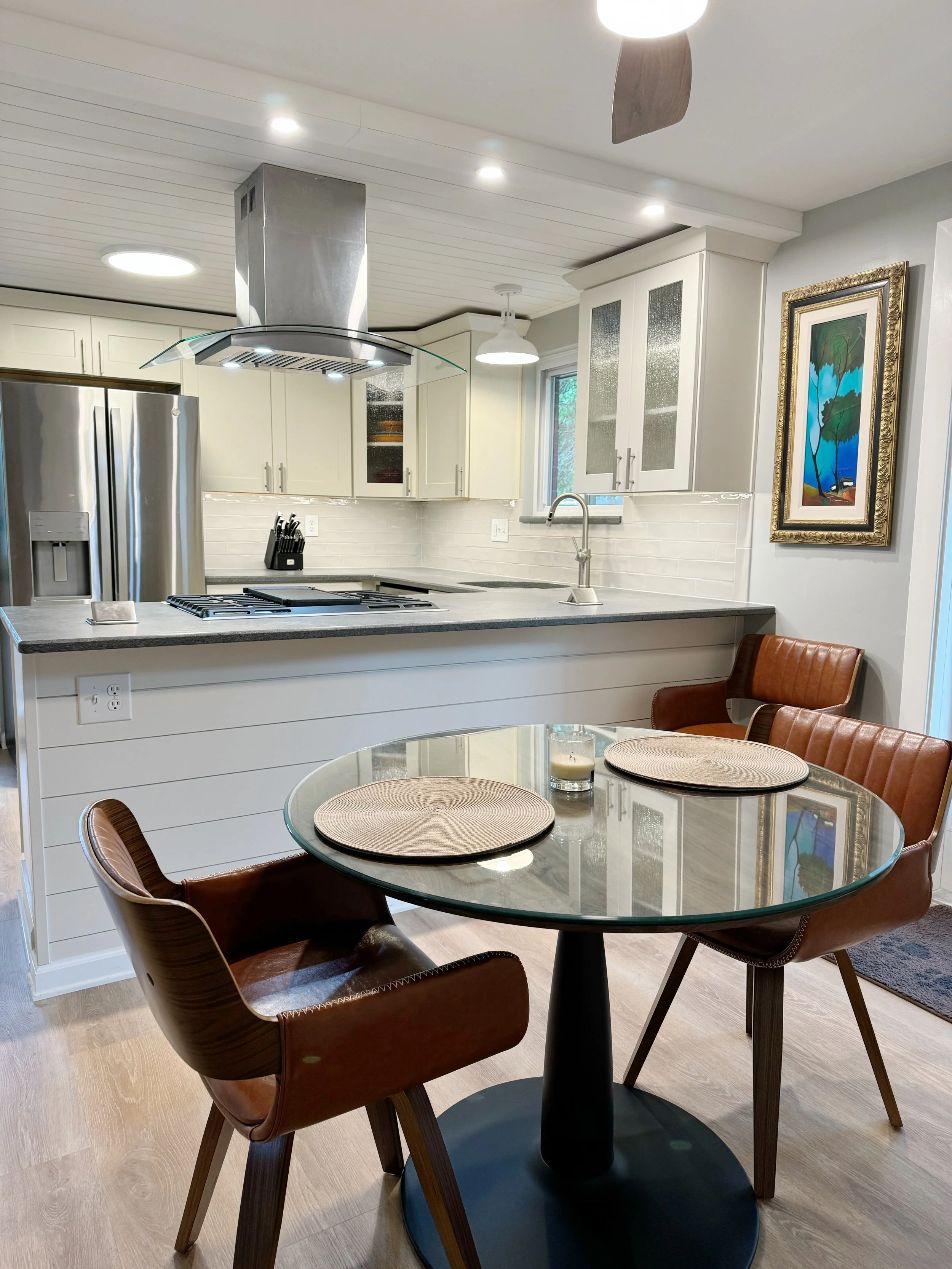 Modern kitchen with white cabinets, stainless steel refrigerator, gas stove, and a round glass dining table with four brown upholstered chairs.