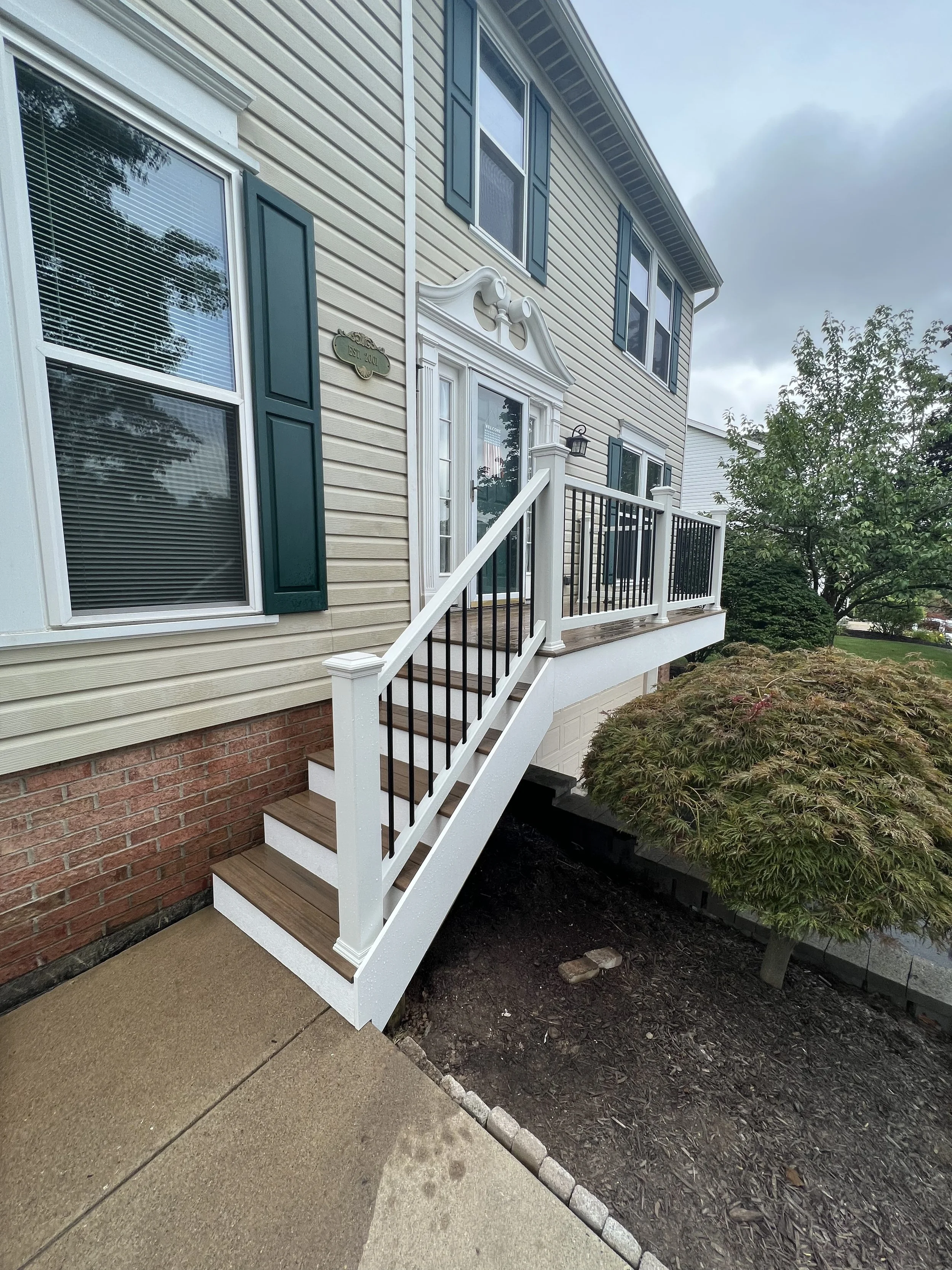 Exterior view of a two-story house with beige siding, white trim, and green shutters. There are stairs leading up to a small balcony with a black and white railing. A gardening bed with a small tree is in the foreground.
