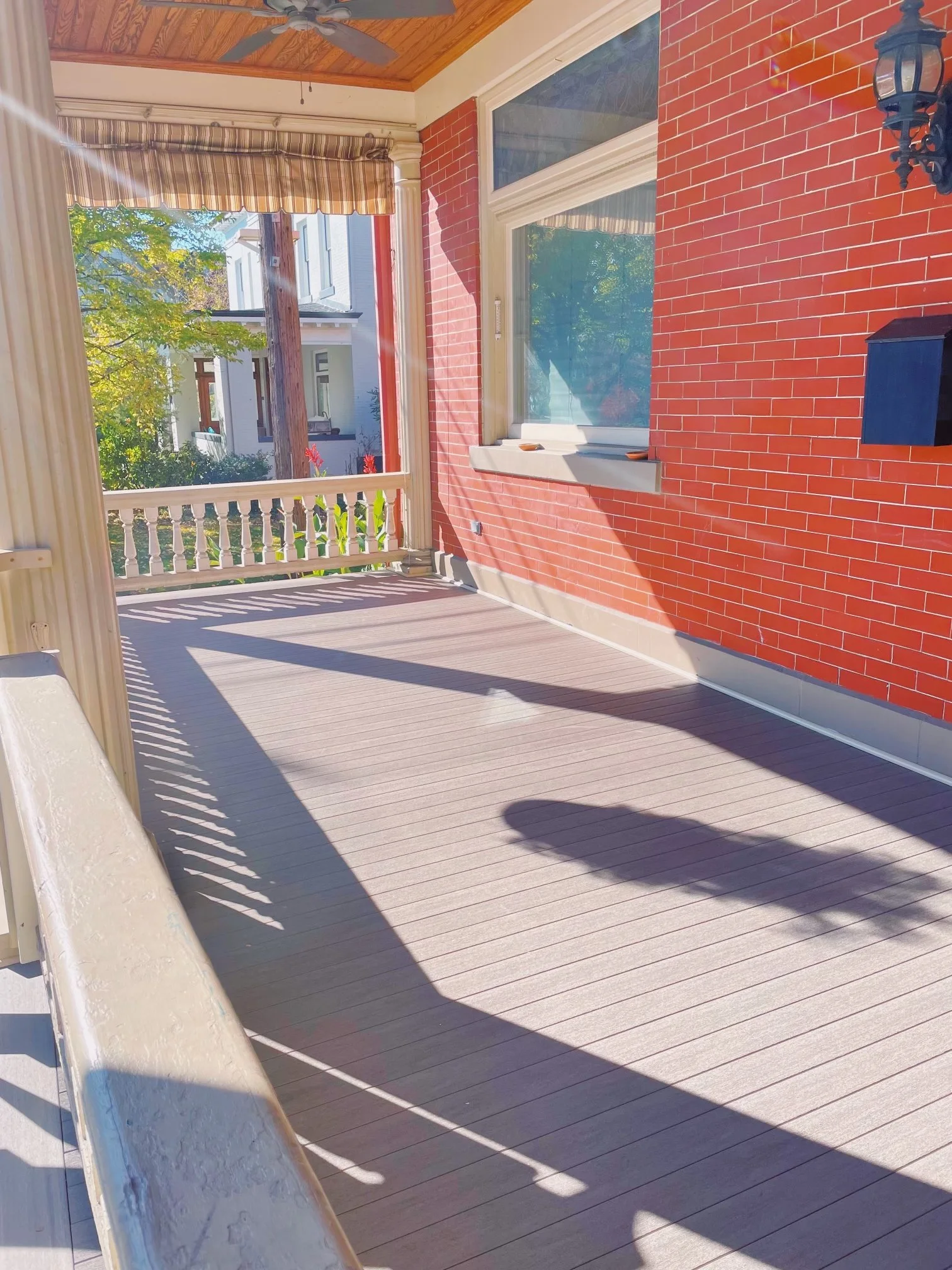 Sunny porch with wooden floor, brick wall, window, railing, hanging lantern, and ceiling fan, with trees visible outside.