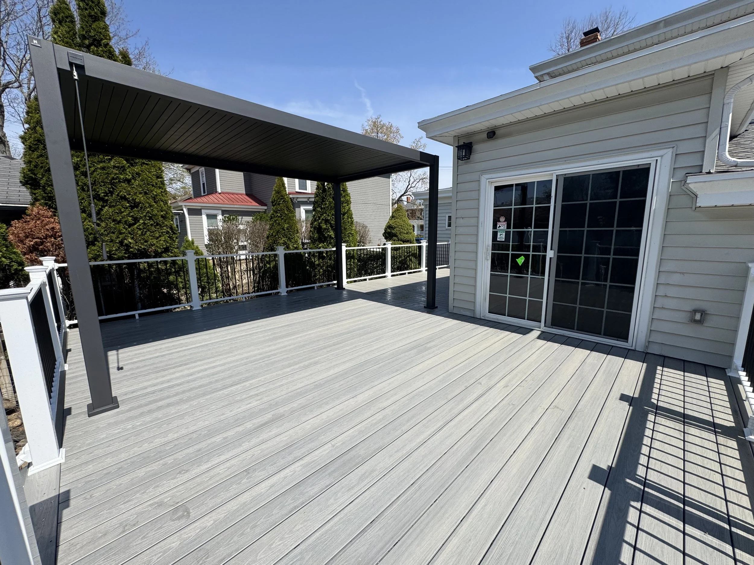 A spacious outdoor deck attached to a house with a glass sliding door and a covered pergola. The deck has white railings and is surrounded by neighboring houses and trees.