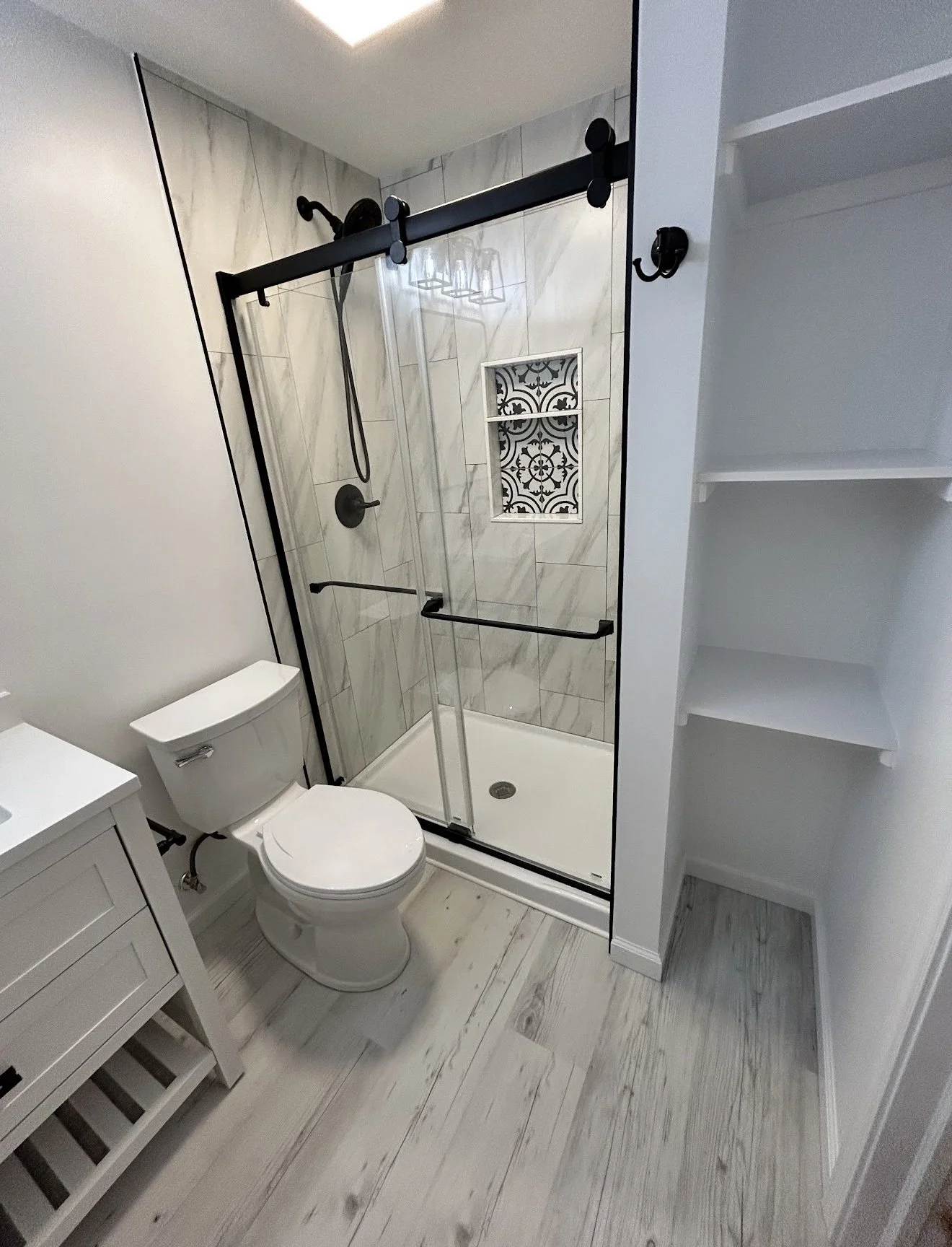 Bathroom with a glass-enclosed shower featuring a decorative tile inset, white toilet, white vanity, and light wood flooring.