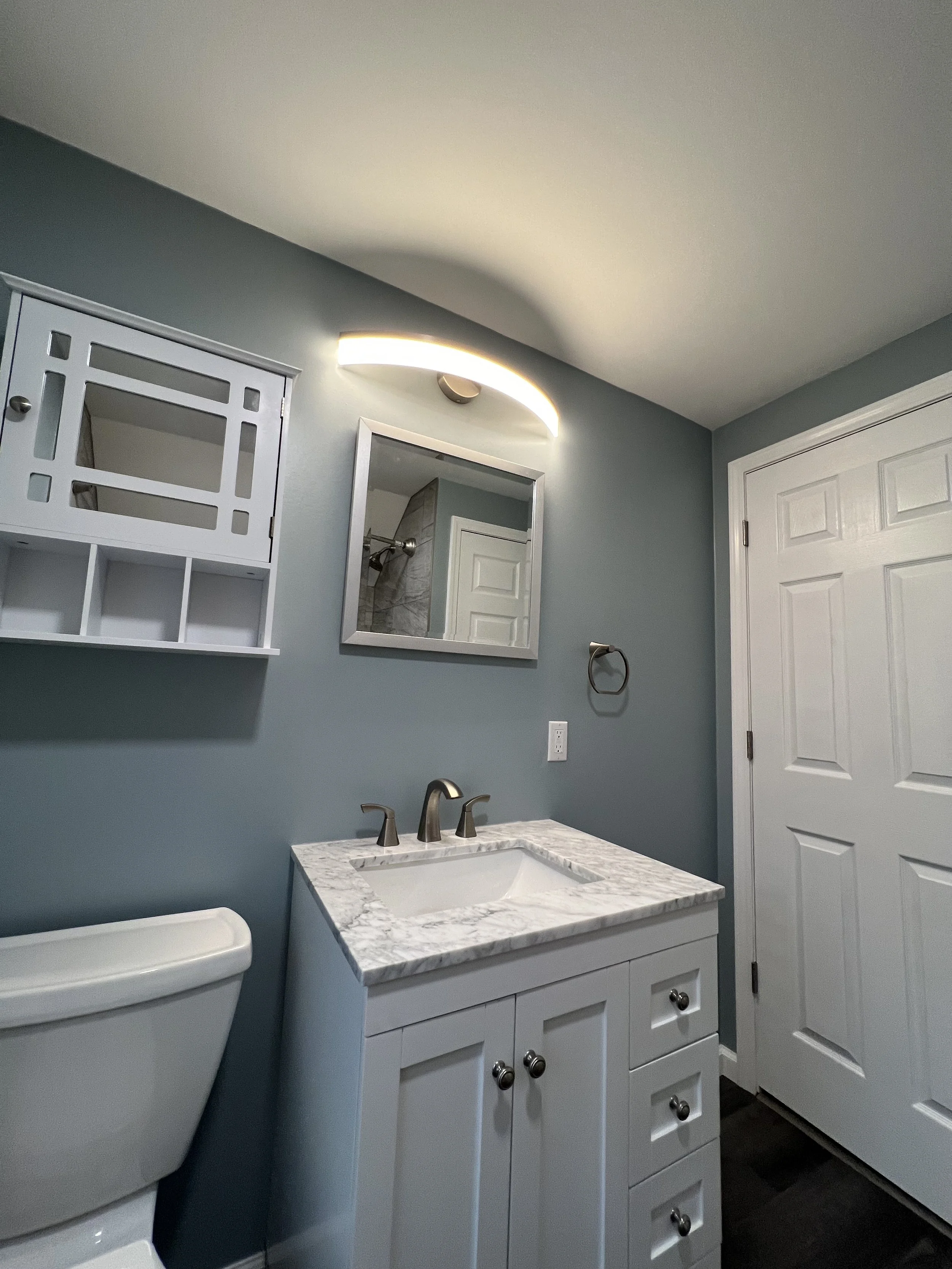 Bathroom with blue walls, white vanity with a marble countertop, small mirror, wall-mounted light fixture, toilet, and a white door.