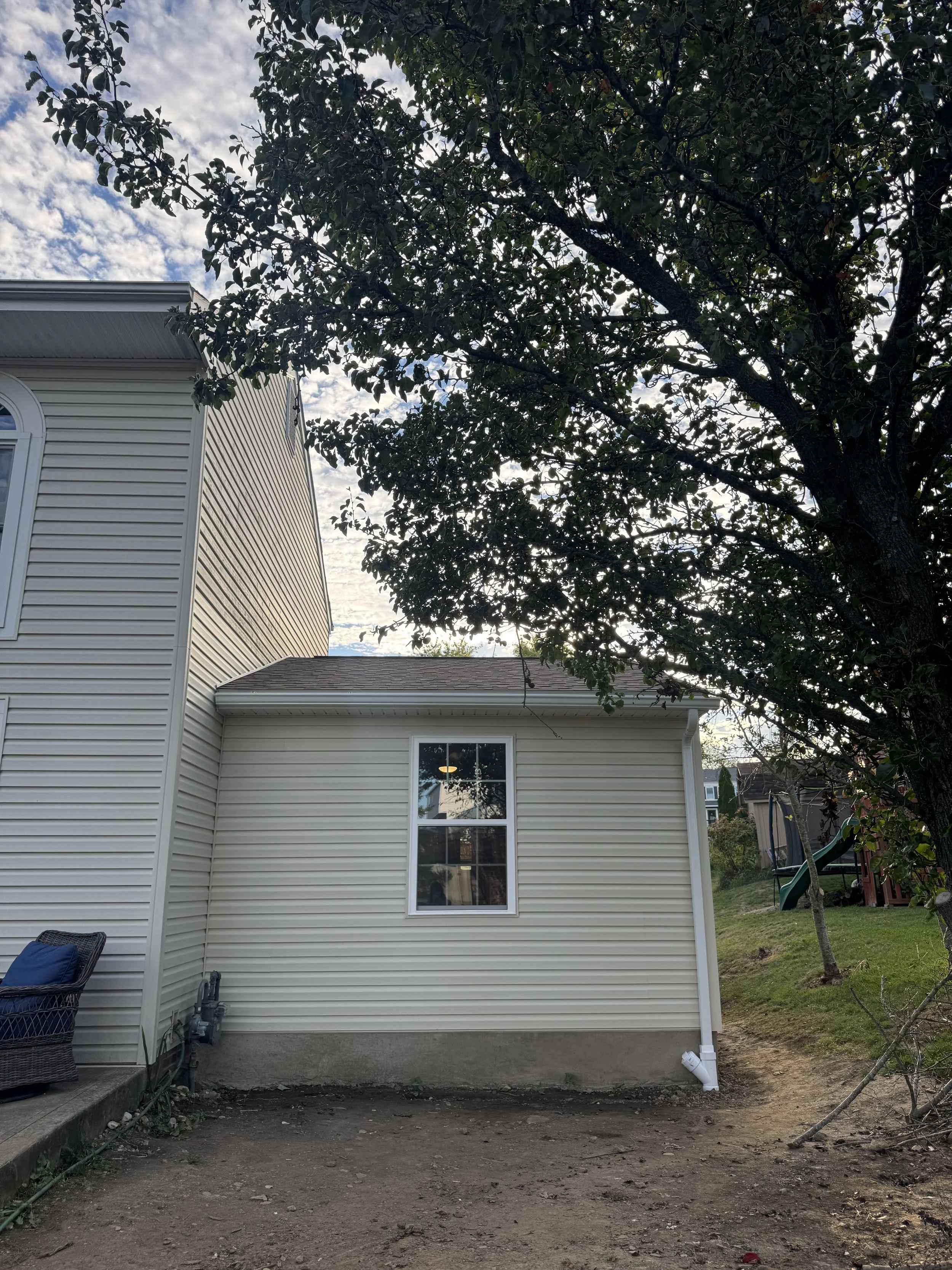 Backyard view of a house with beige siding, a window, a tree, and a dirt yard.