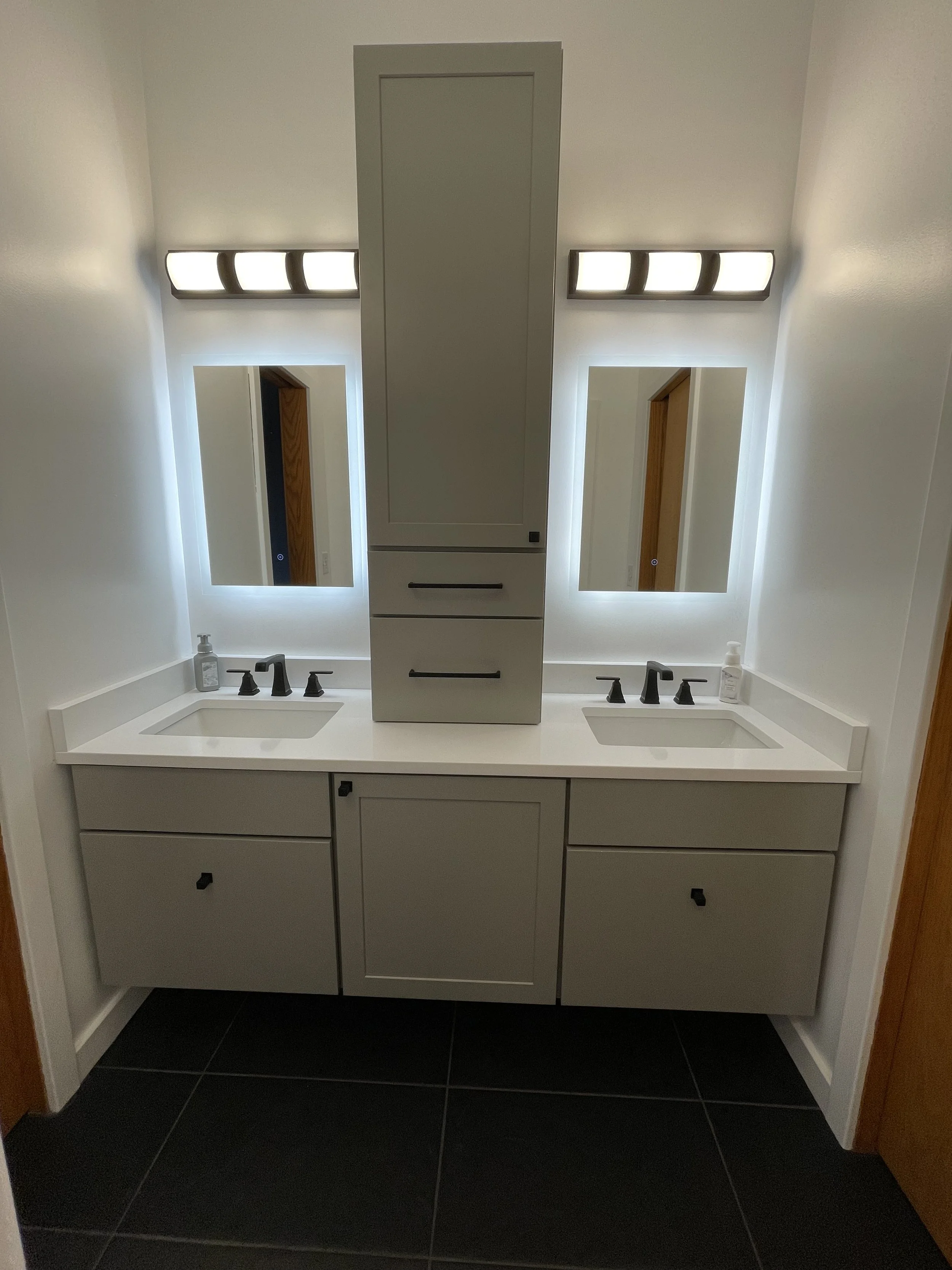 A modern bathroom vanity with two sinks, illuminated mirrors, a central cabinet with drawers, and black fixtures, on a black tiled floor.