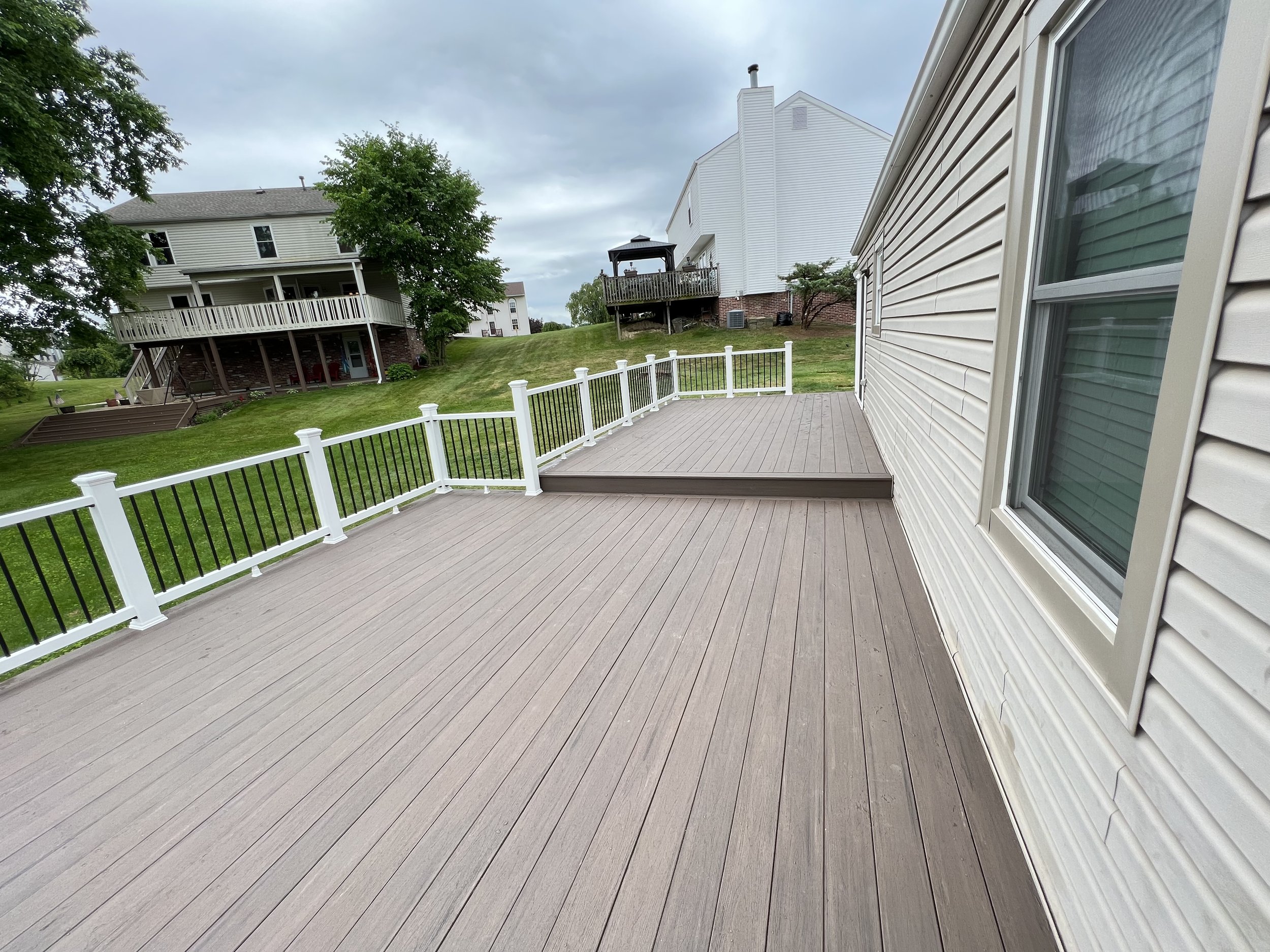 A spacious outdoor deck with beige-colored flooring and white railing, attached to a house with beige siding, overlooking a grassy yard and neighboring houses with decks and trees under an overcast sky.