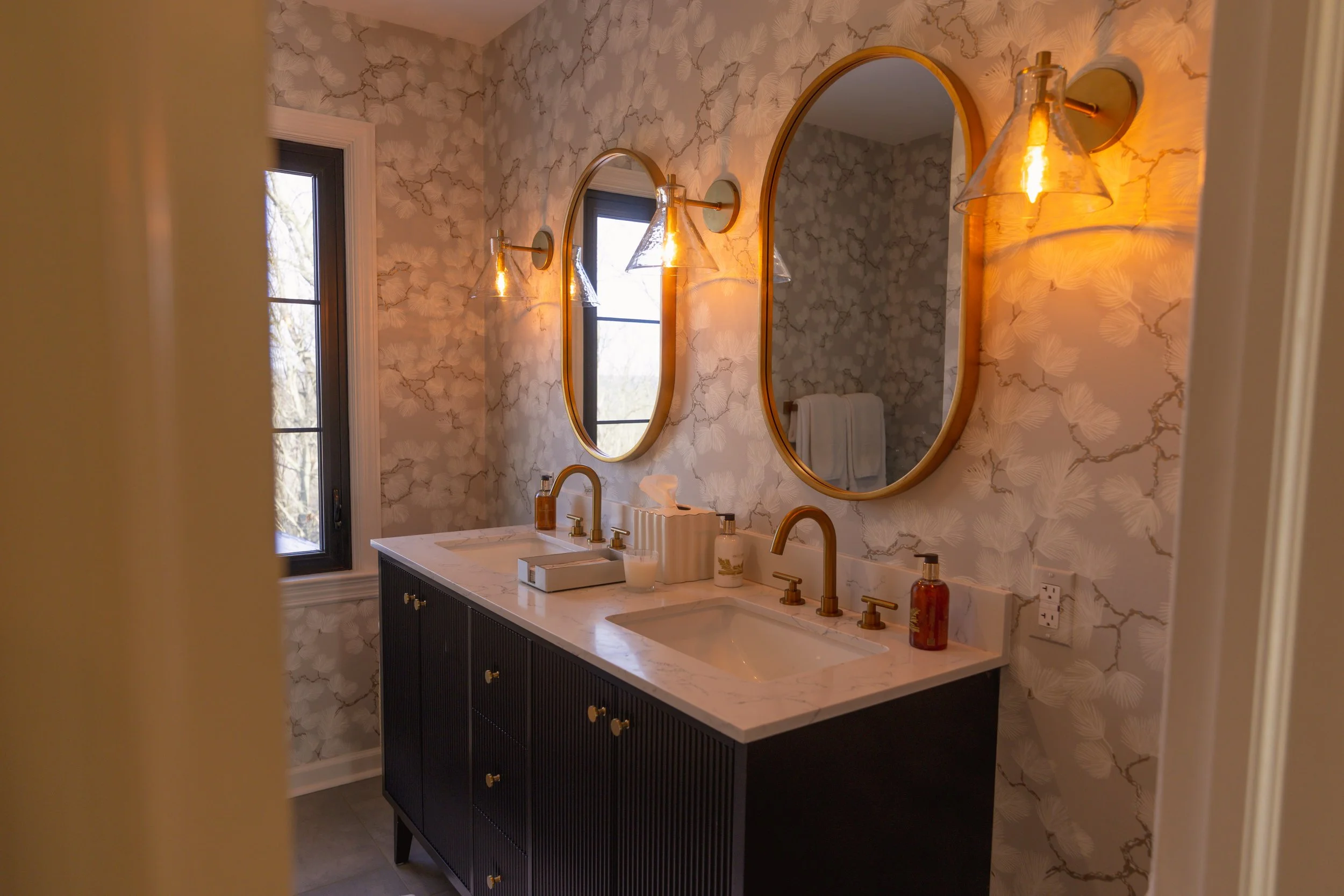 A modern bathroom with a black double vanity with gold knobs, a white marble countertop, two oval gold-framed mirrors, gold wall-mounted light fixtures with glass shades, a window with black trim, and wallpaper with a delicate floral pattern.