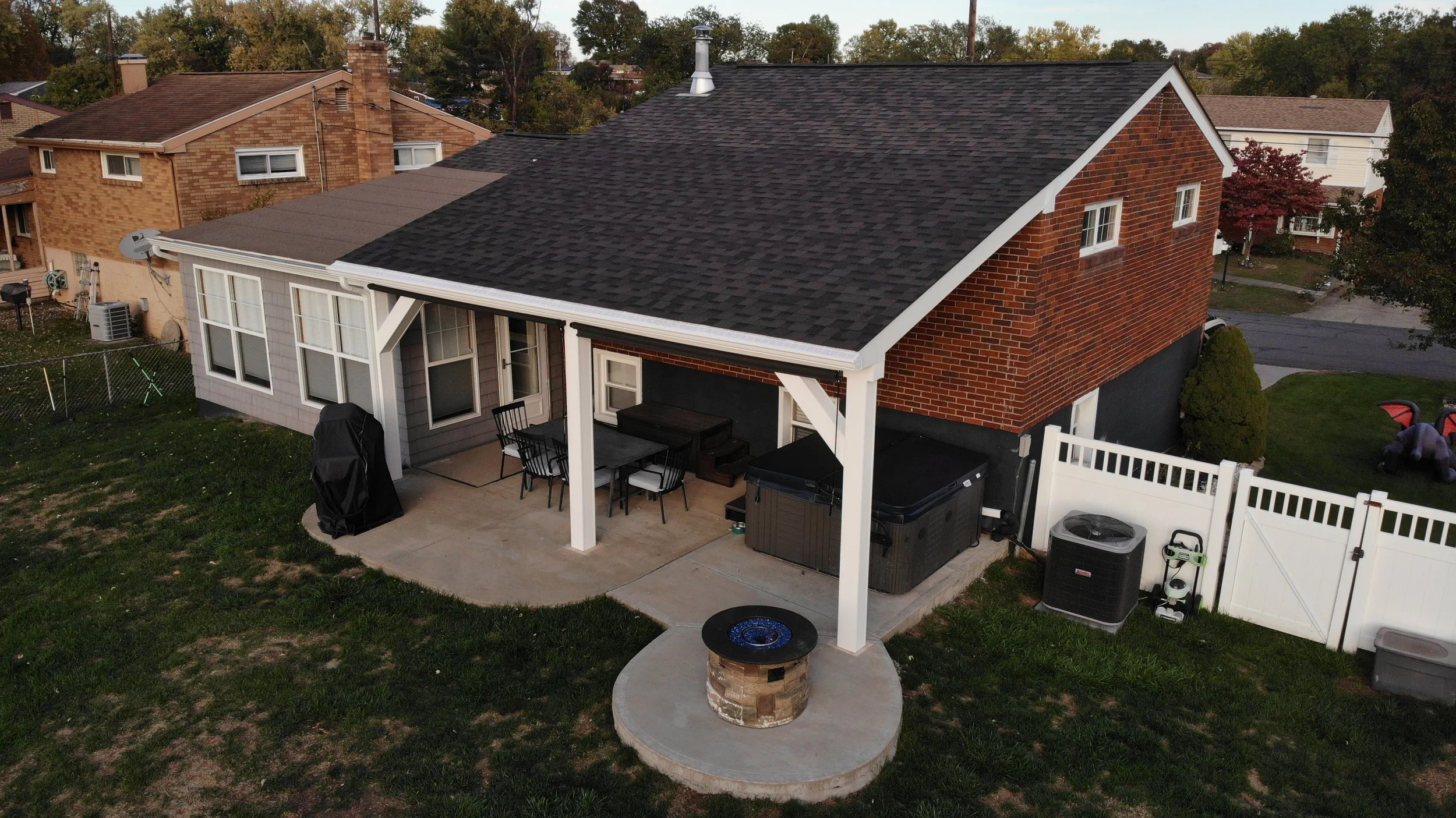 A backyard patio area with outdoor furniture, a grill, a fire pit, and a hot tub, enclosed by a white fence behind a brick house with a covered porch.