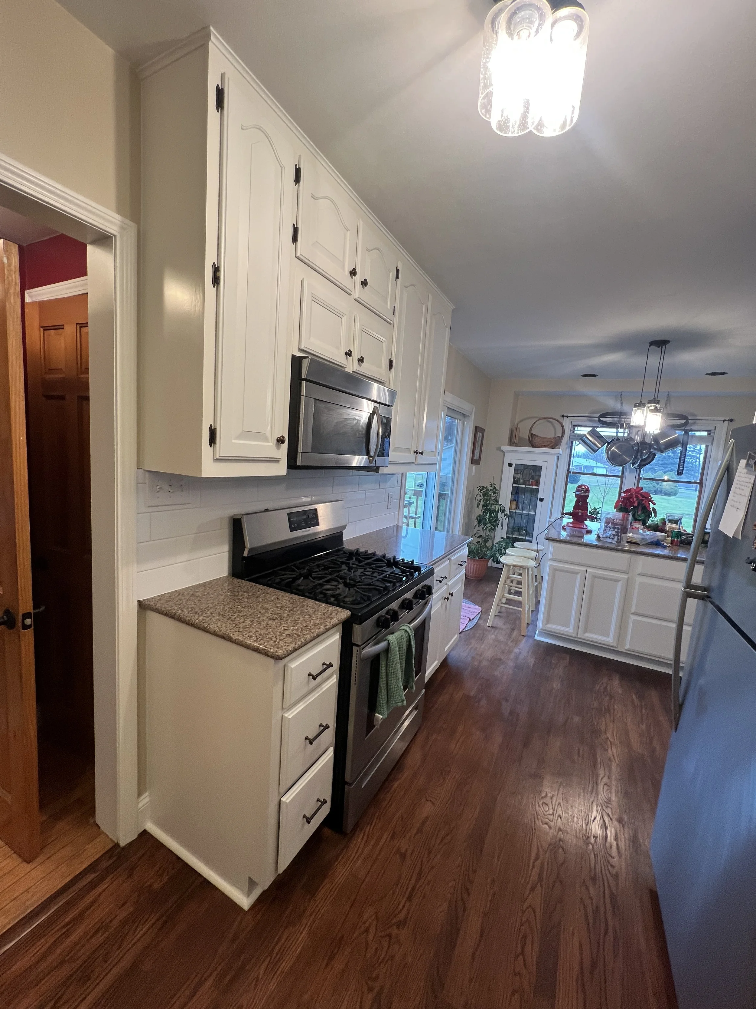 Kitchen with white cabinets, granite countertops, stainless steel stove and microwave, wooden flooring, a refrigerator, and a dining area with a window and hanging lights.