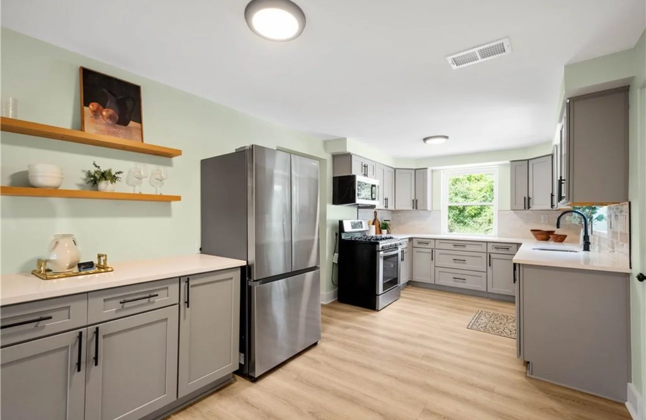 Modern L-shaped kitchen with light gray cabinets, stainless steel refrigerator, black stove and microwave, light-colored countertops, a window with greenery outside, and wooden open shelves with decor on the wall.