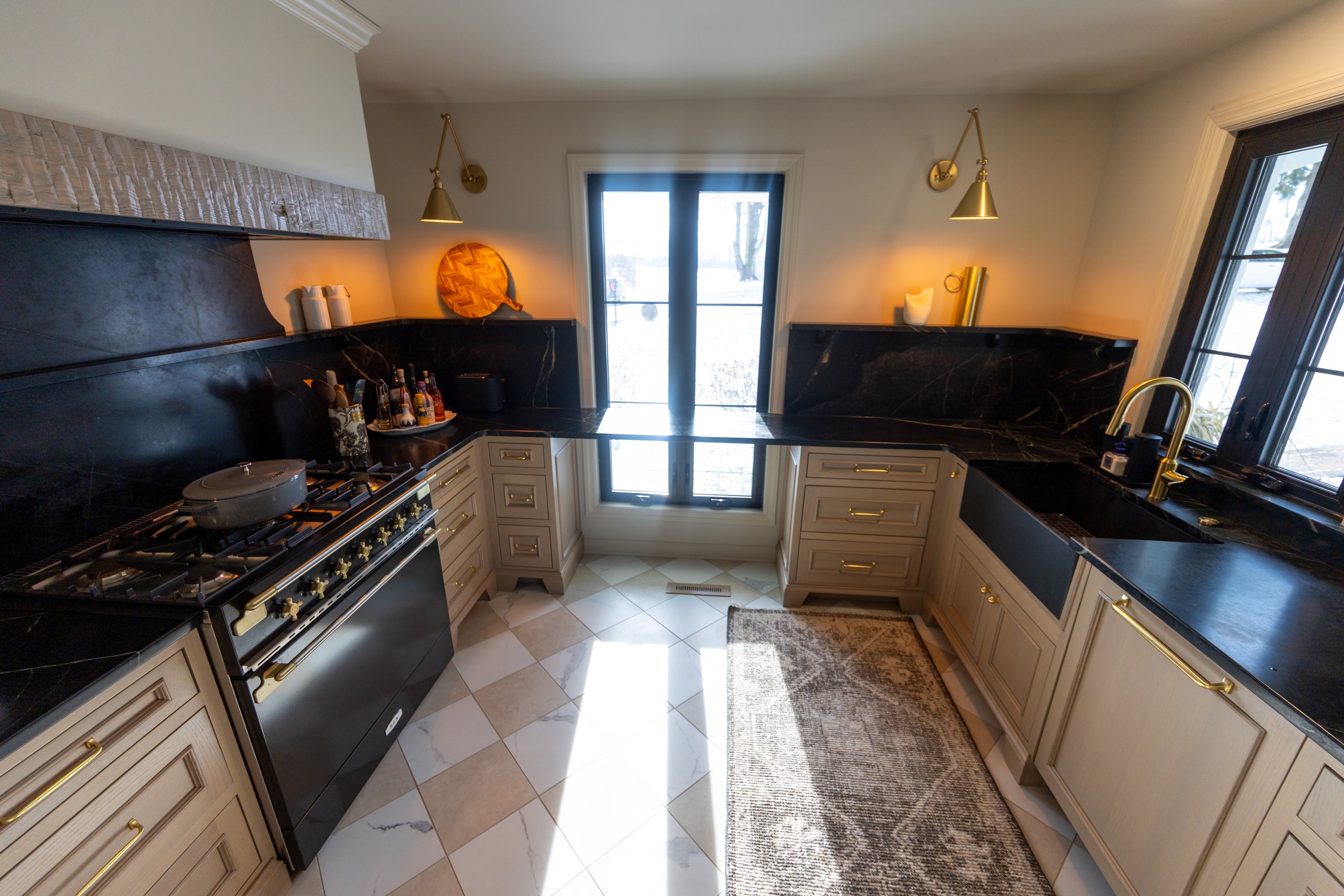 Kitchen with black marble countertops, beige cabinets with gold handles, a black oven, and a black sink, with a window and sliding glass door letting in natural light, and decorative wall lights and accents.