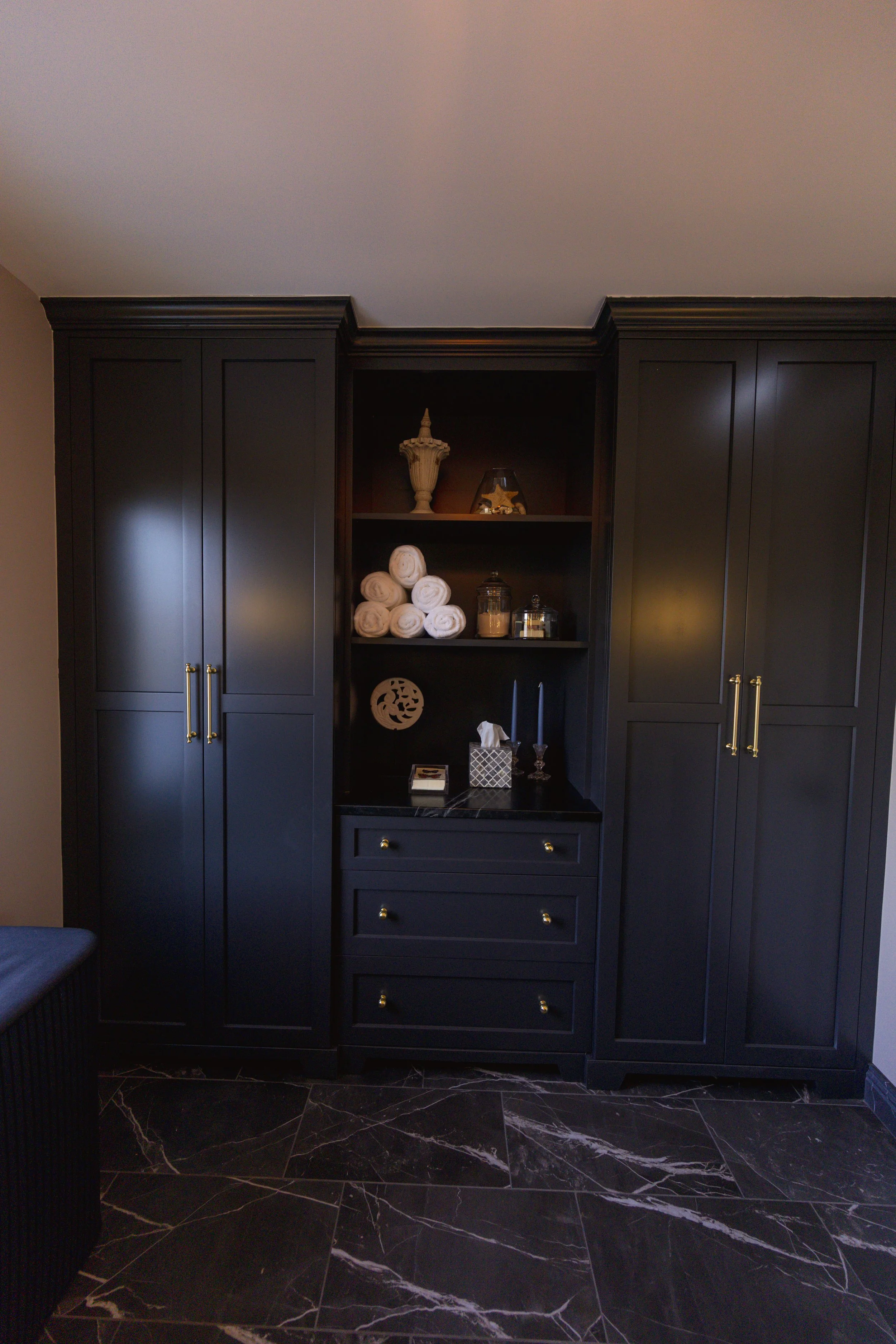 Black built-in cabinet with closed doors on the left and right, and open shelves in the center displaying rolled towels, decorative jars, and ornaments, with black marble countertop and black marble floor.