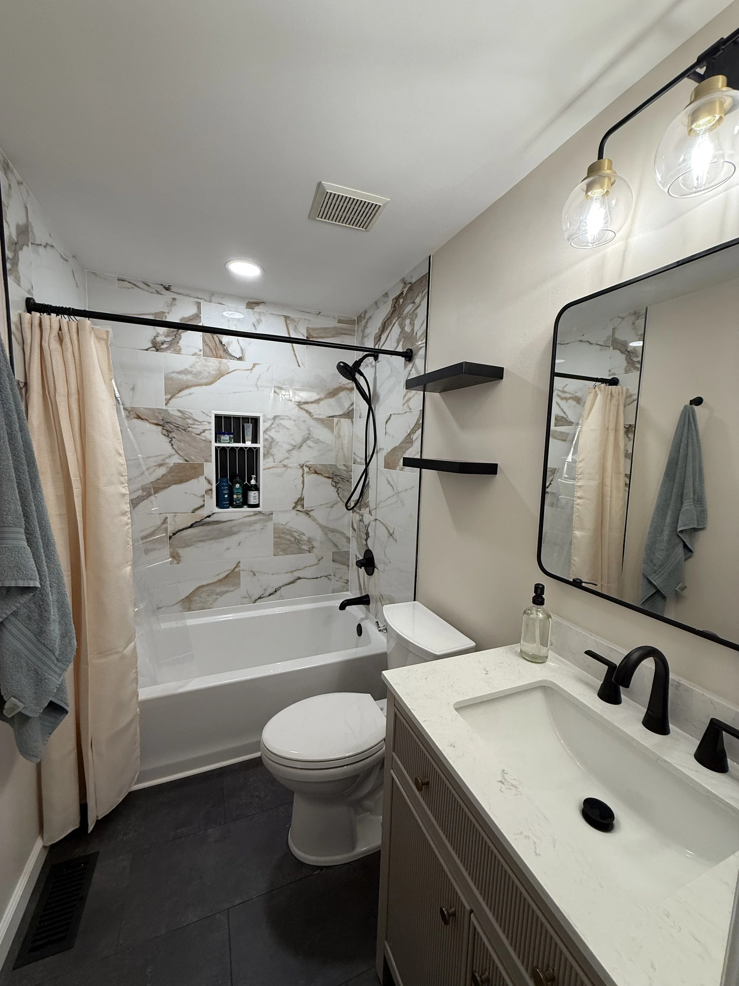 Bathroom with a bathtub and shower combination with marble tile walls, a toilet, a vanity with a sink, a mirror, and wall-mounted shelves. The shower has a black showerhead and a built-in shelf with toiletries. Towel hooks or bars hold towels, and a 
