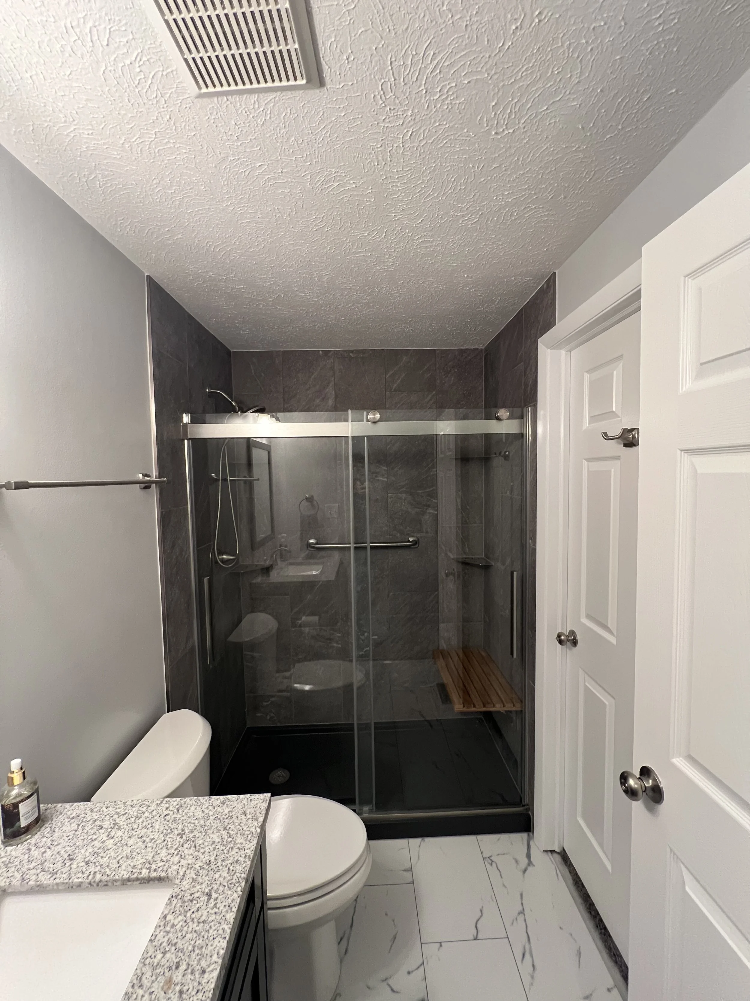 A modern bathroom with a black tile shower enclosure, white toilet, granite countertop, and white doors. The shower has a glass door with a metal handle, a wooden shower bench, and a wall-mounted mirror.