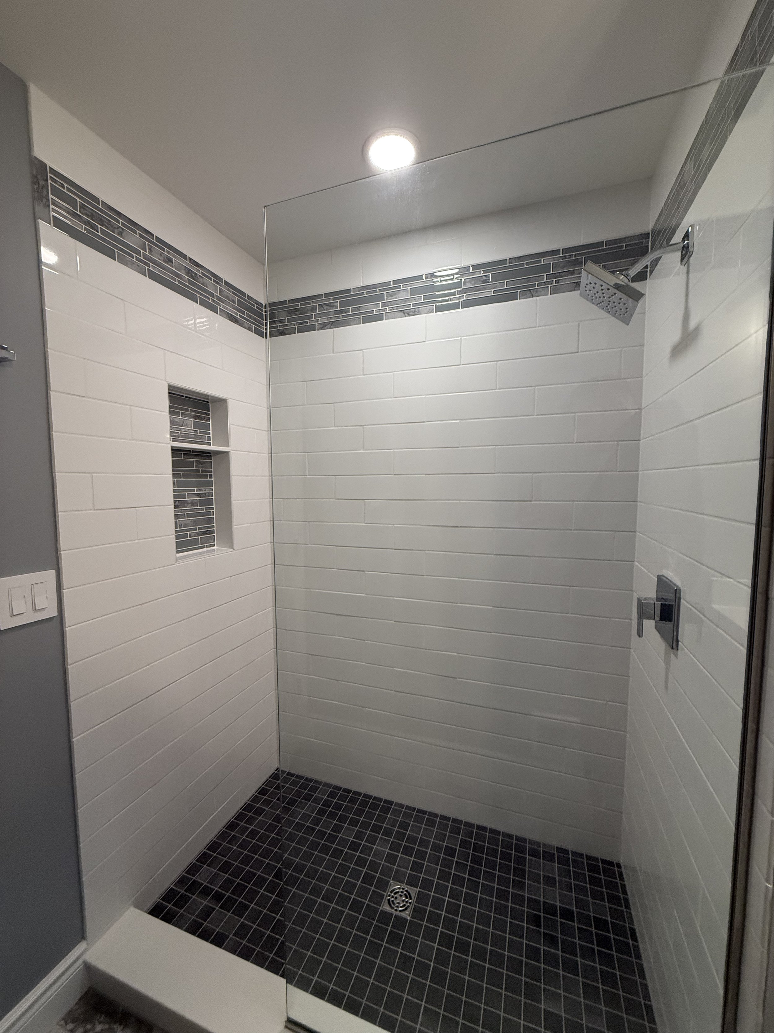 Empty walk-in shower with white tiled walls, black mosaic floor tiles, a small built-in wall niche with black mosaic tiles, a silver overhead showerhead, a control knob, and a glass partition.