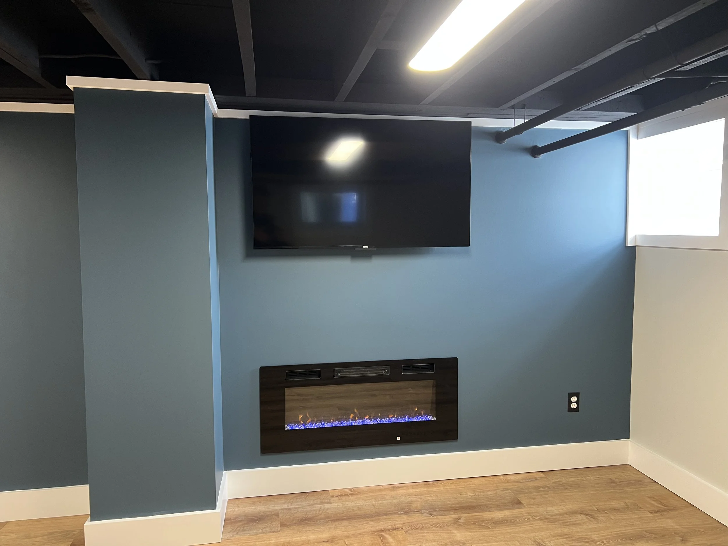 Modern living room wall with mounted flat screen TV and electric fireplace, painted in blue tones, with a window on the right side.