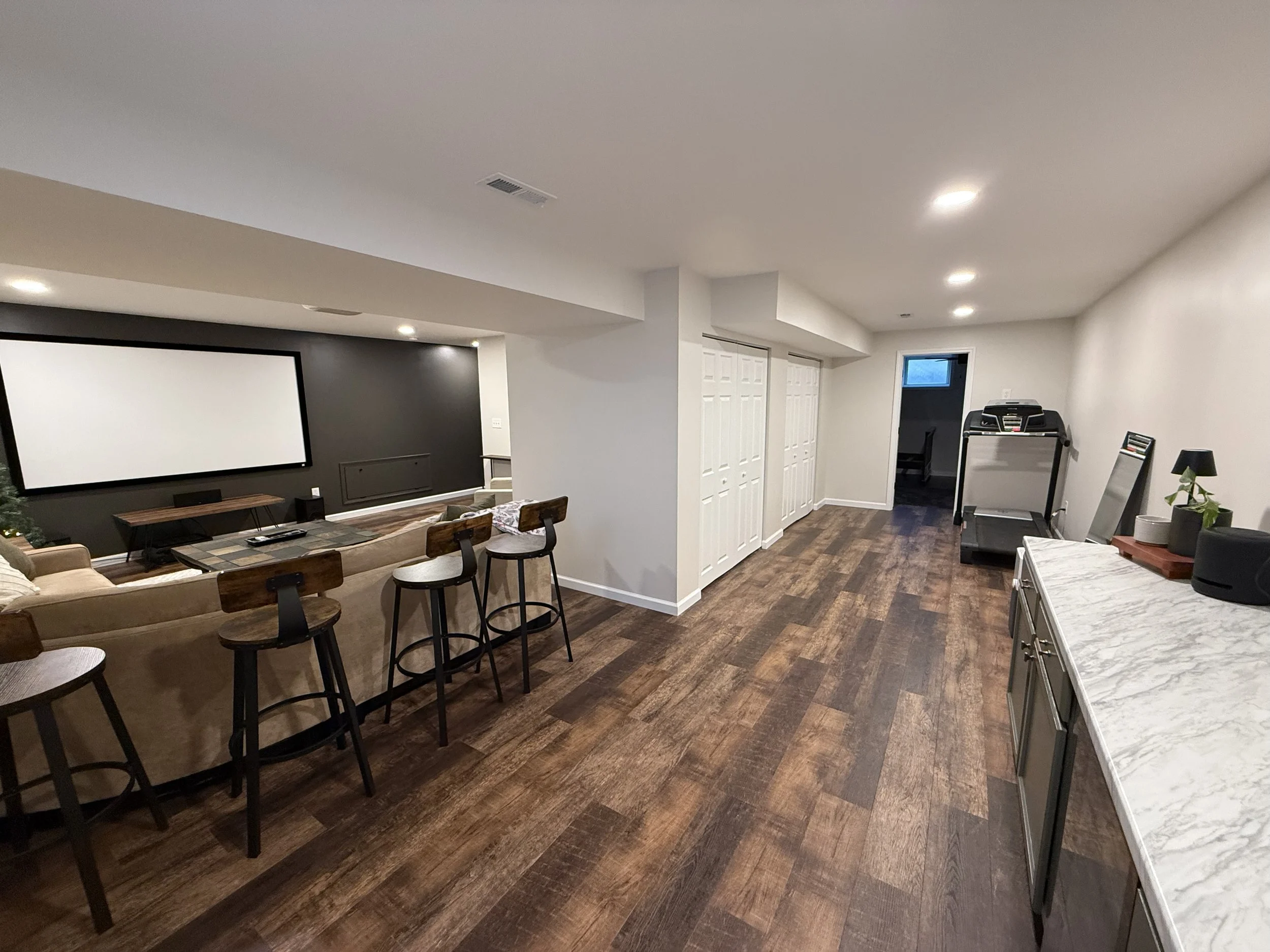 A basement entertainment room with a large white screen, a dark accent wall, a beige sectional sofa, a TV stand, a countertop with barstools, a treadmill, and a small table with plants and a speaker.