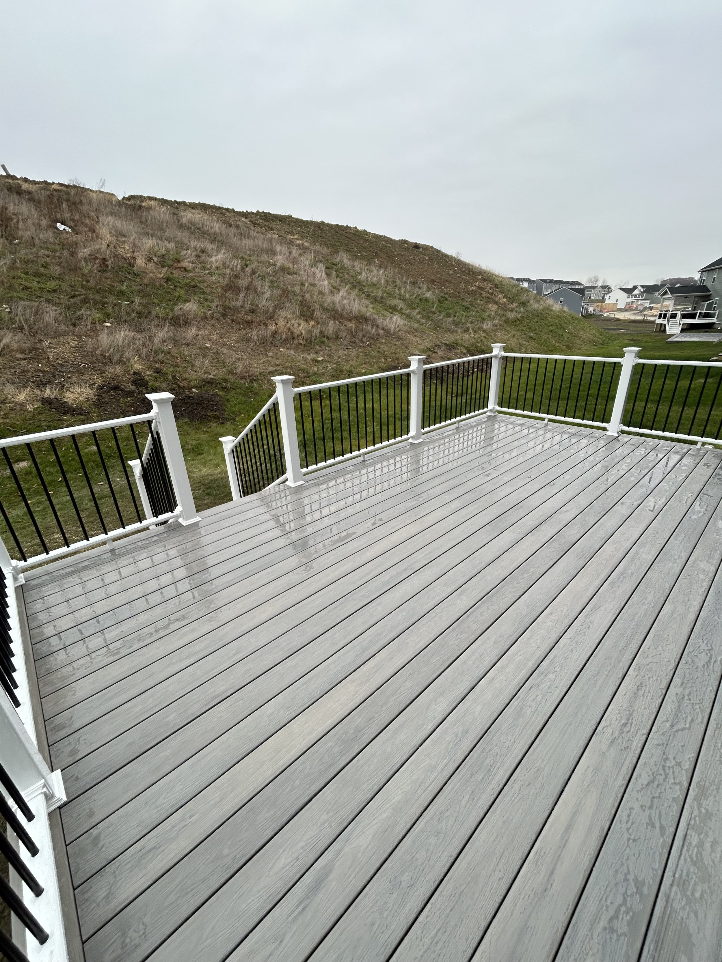 Wet wooden deck with white and black railing overlooking a hillside and neighboring houses on a cloudy day.