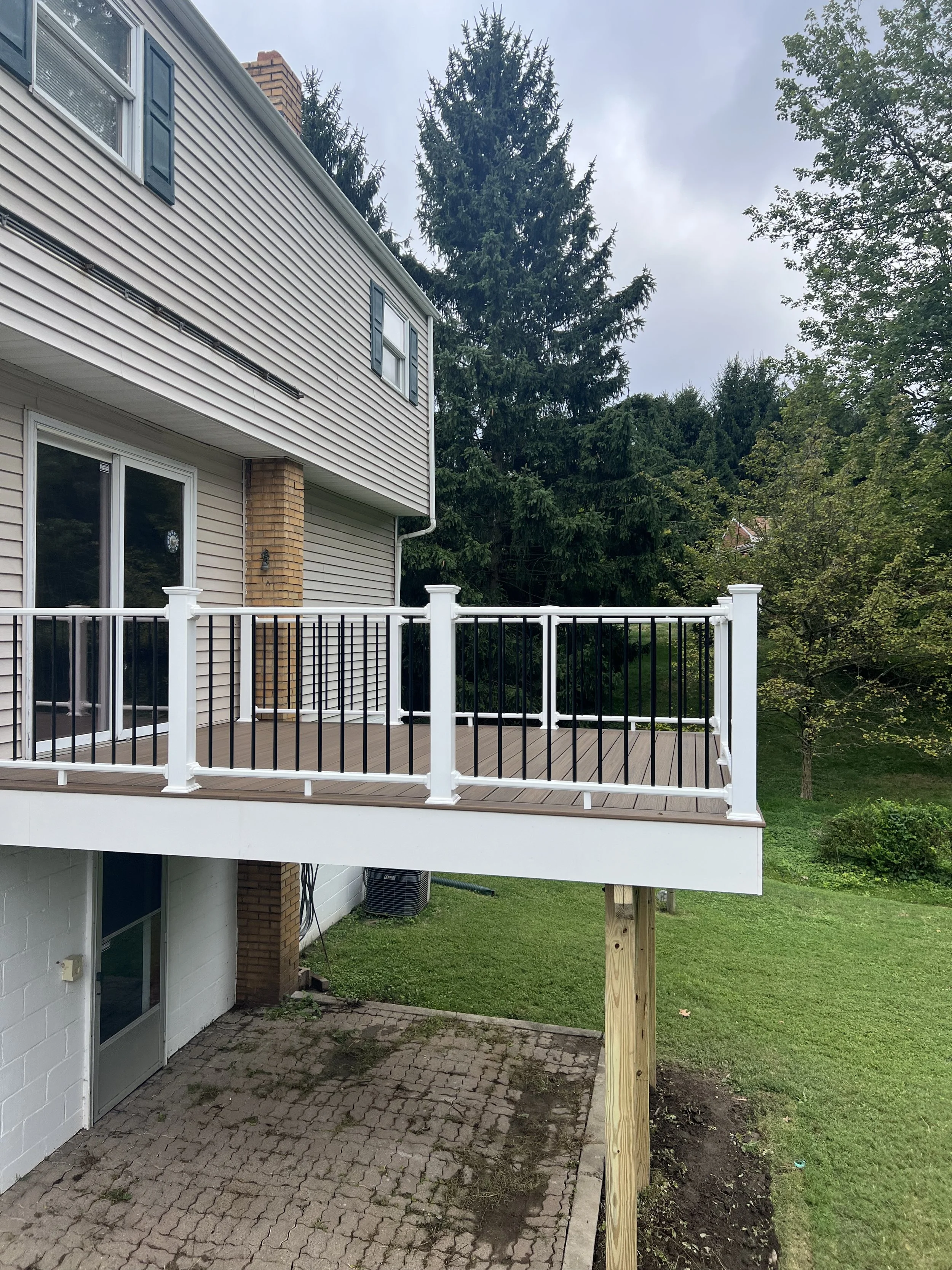 A residential house with a balcony, white and black railing, brown wooden floor, sliding glass door, brick chimney, beige siding, green lawn, and trees in the background under a cloudy sky.