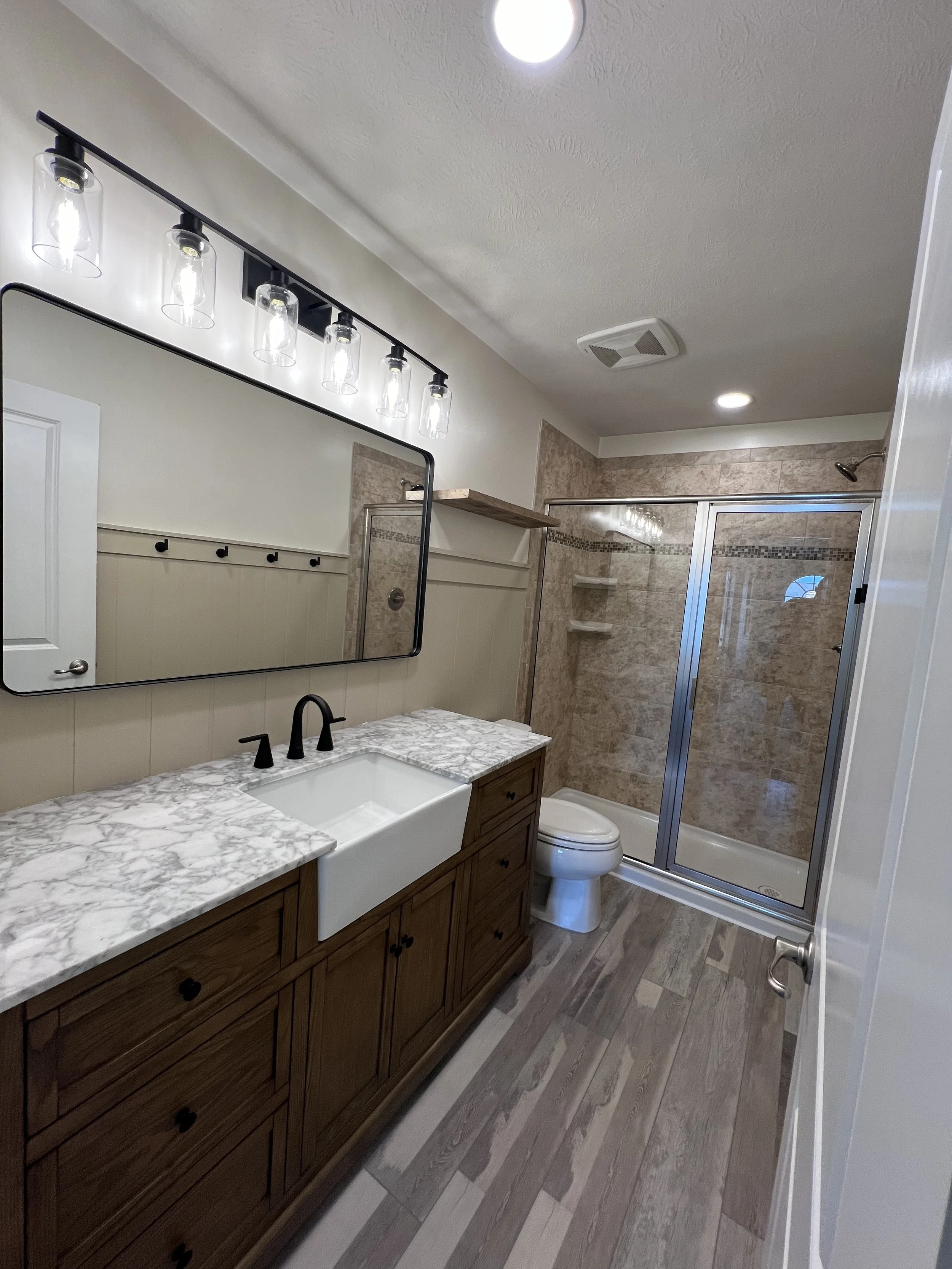 A modern bathroom with a large mirror, farmhouse sink, marble countertop, wooden cabinet, enclosed shower with beige tiles, and a toilet.